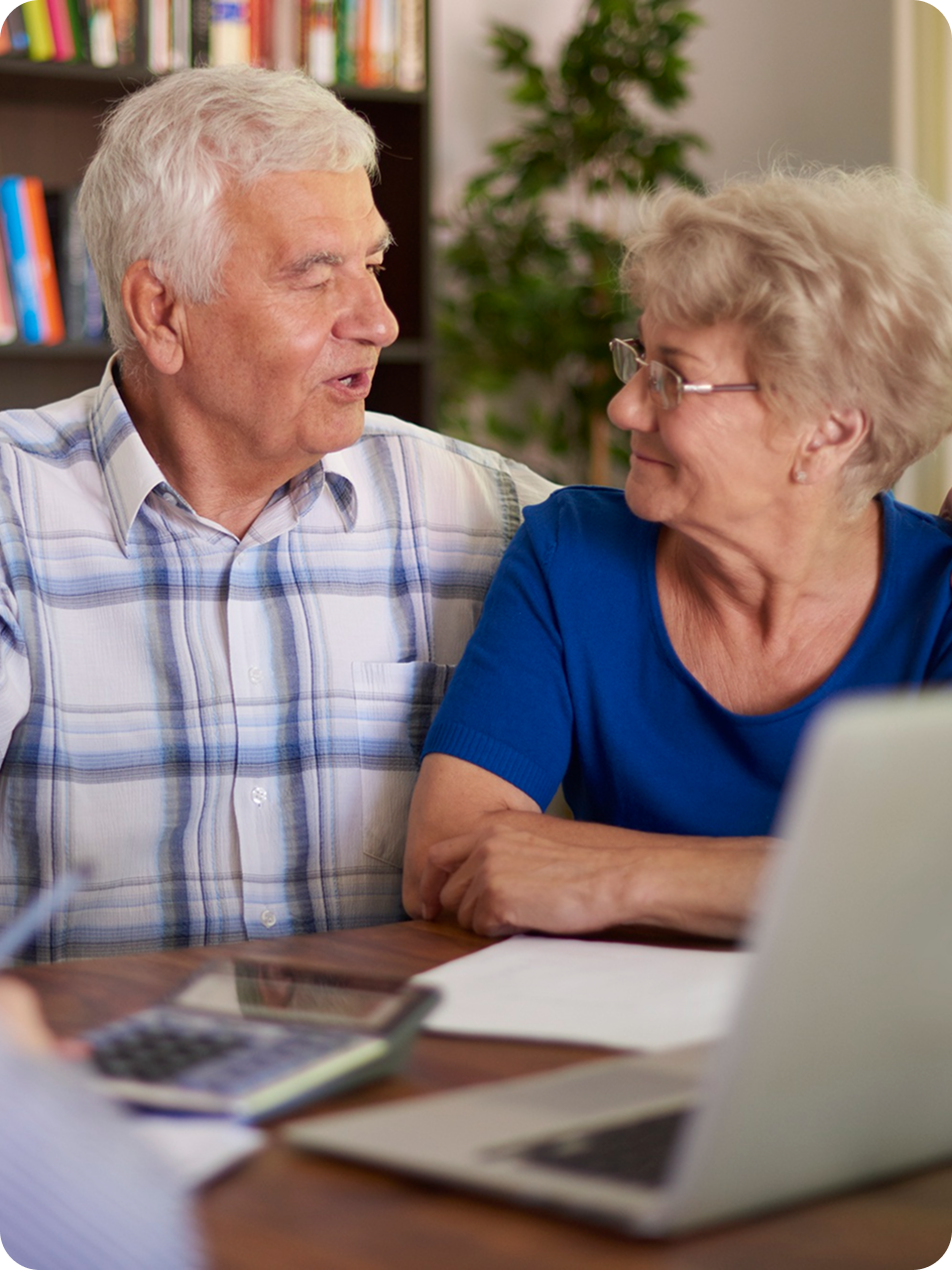 An elderly man and woman sitting together at a table, engaged in conversation, with a laptop, calculator, and papers in front of them.