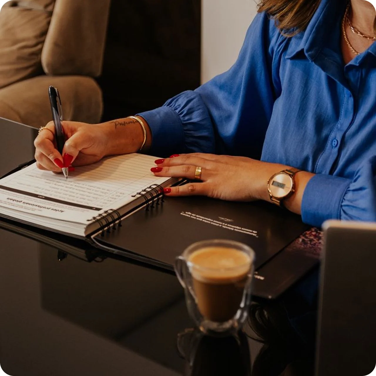 A woman wearing a blue shirt and gold watch is writing in a spiral notebook with a black pen, with her left hand resting on the open notebook. The notebook is on a black table, and there is a small glass cup of espresso coffee in the foreground. A brown sofa is visible in the background.