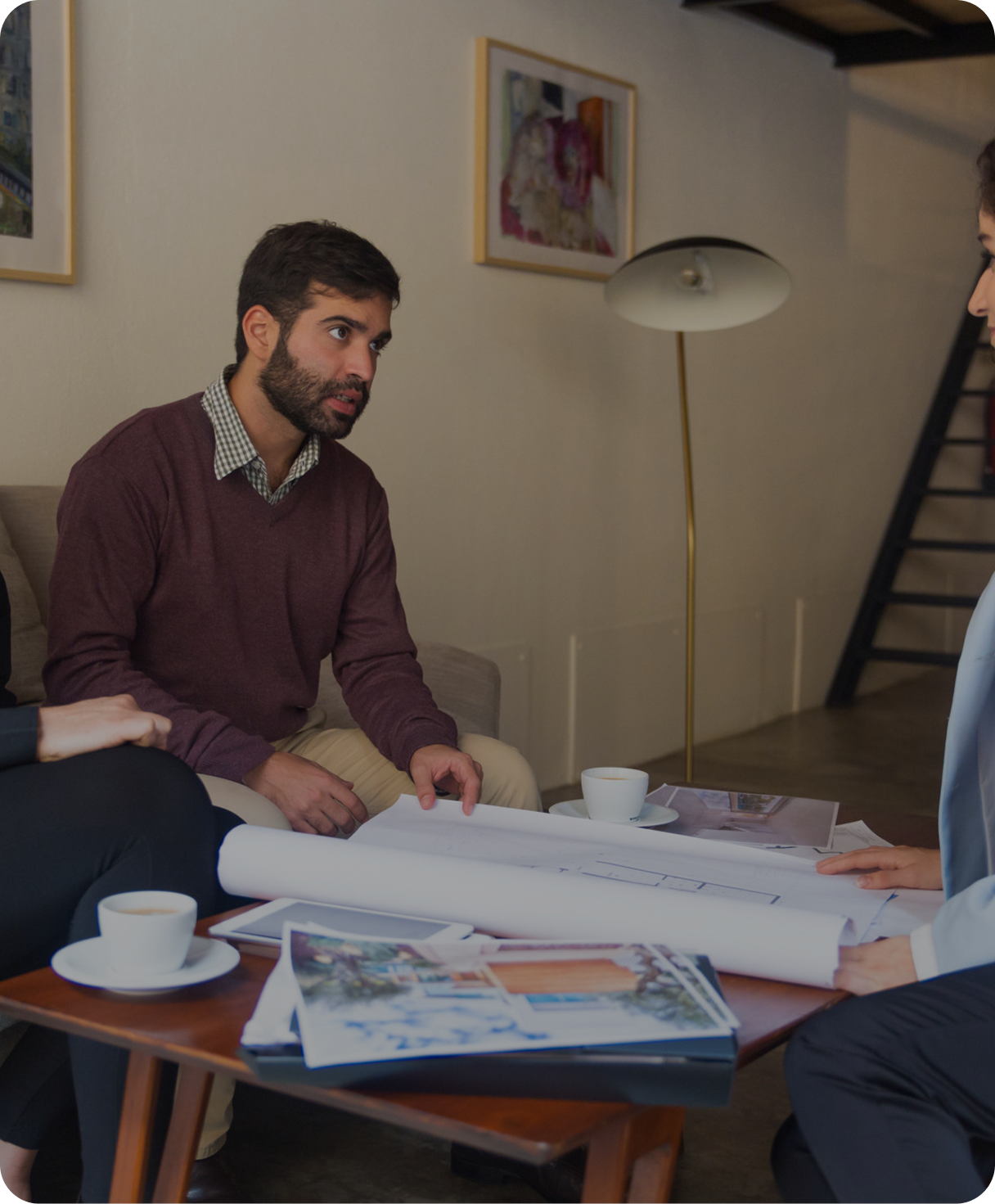 A man with brown hair and beard wearing a maroon sweater over a checkered shirt is sitting on a beige couch, discussing plans with a woman across a table. The table has rolled-up blueprints, photographs, and cups of coffee. A lamp and framed artwork are visible in the background.