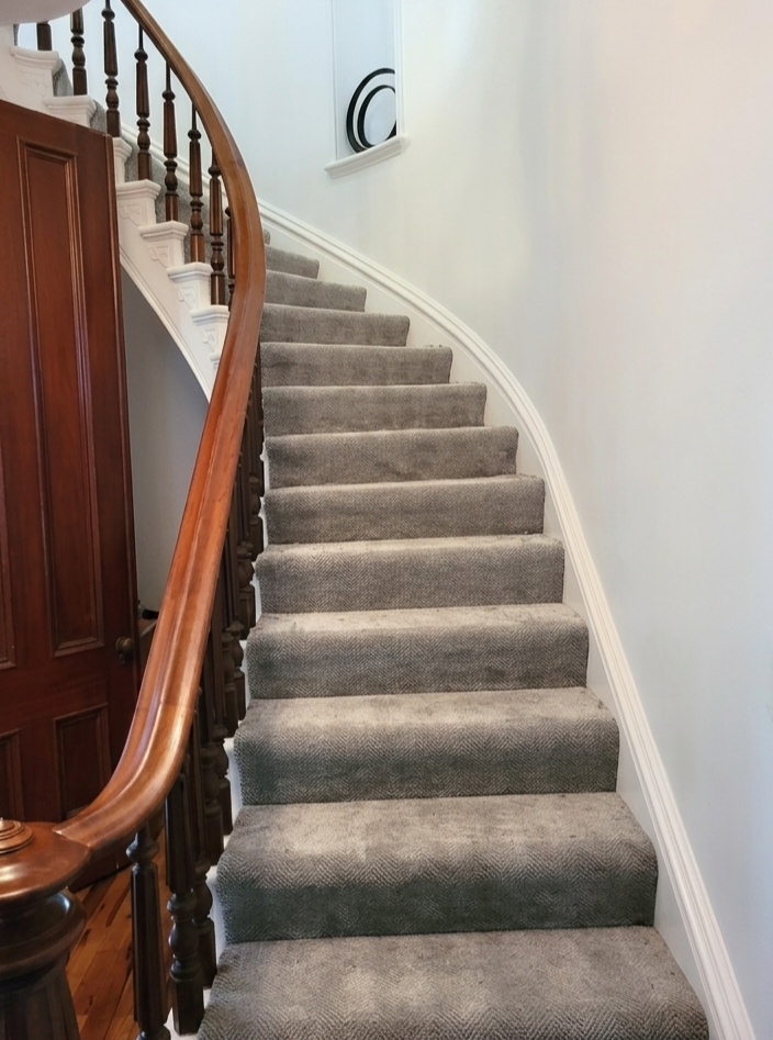 Carpeted staircase with wooden handrail and white wall on the right, curved and leading upward.