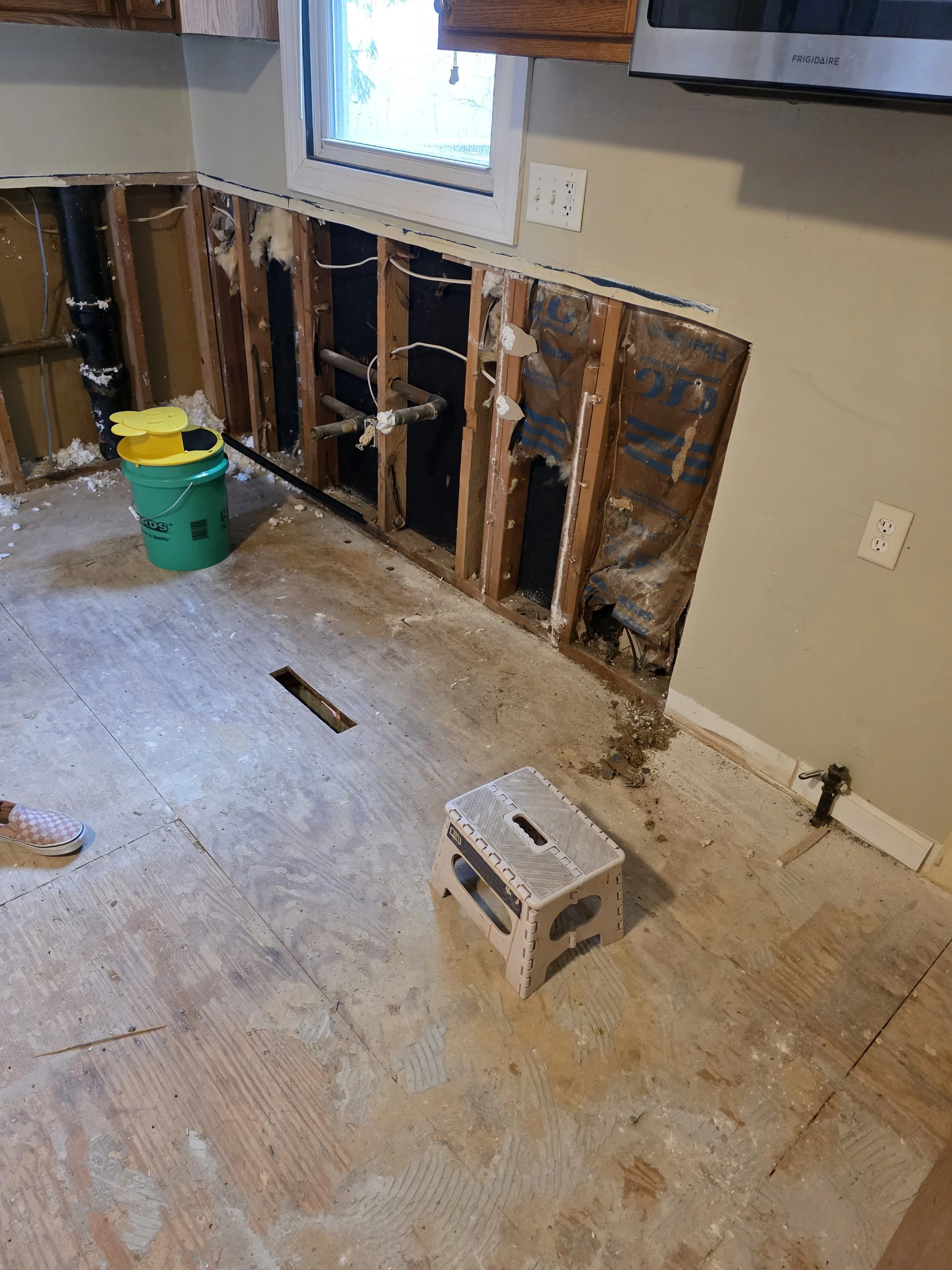 A room under renovation with exposed wall studs, pipes, and insulation. The floor is unfinished with visible dirt and debris. A small plastic step stool is in the center of the room. A bucket with a yellow lid is on the floor near the wall. There is a window with a white frame and a microwave mounted above the counter.