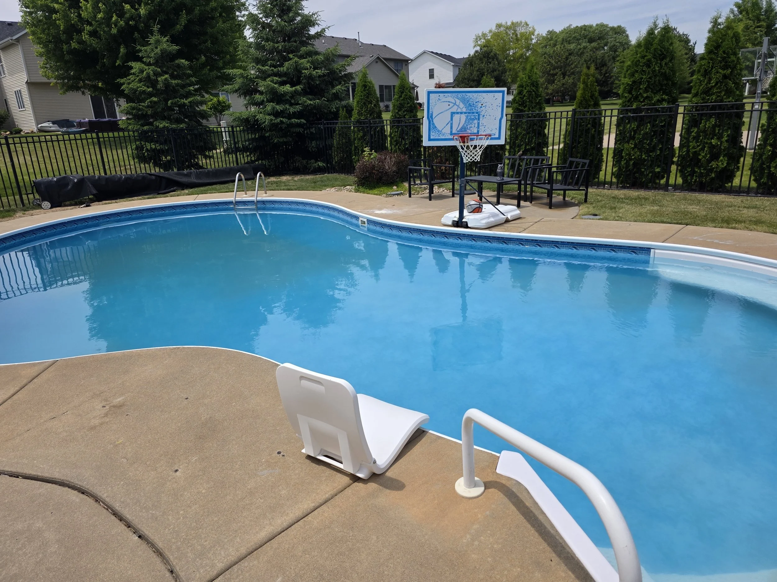 Residential backyard with a swimming pool, basketball hoop, and trees along the fence line.
