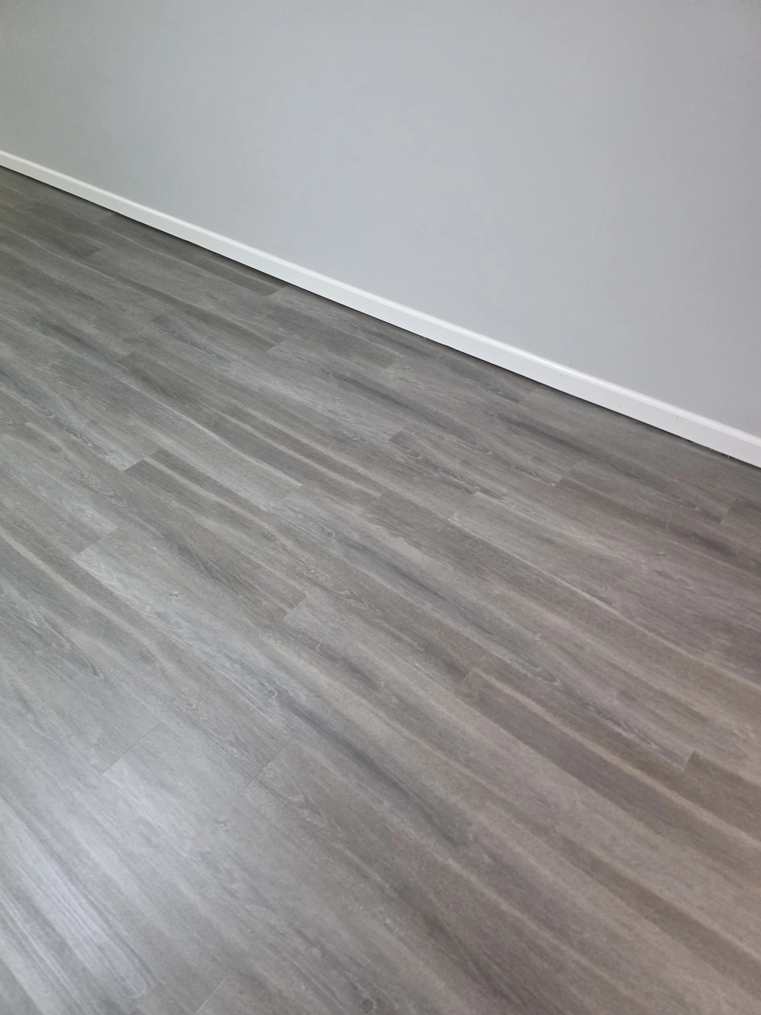 Close-up of a gray painted wall with a white baseboard and gray wood-look flooring.
