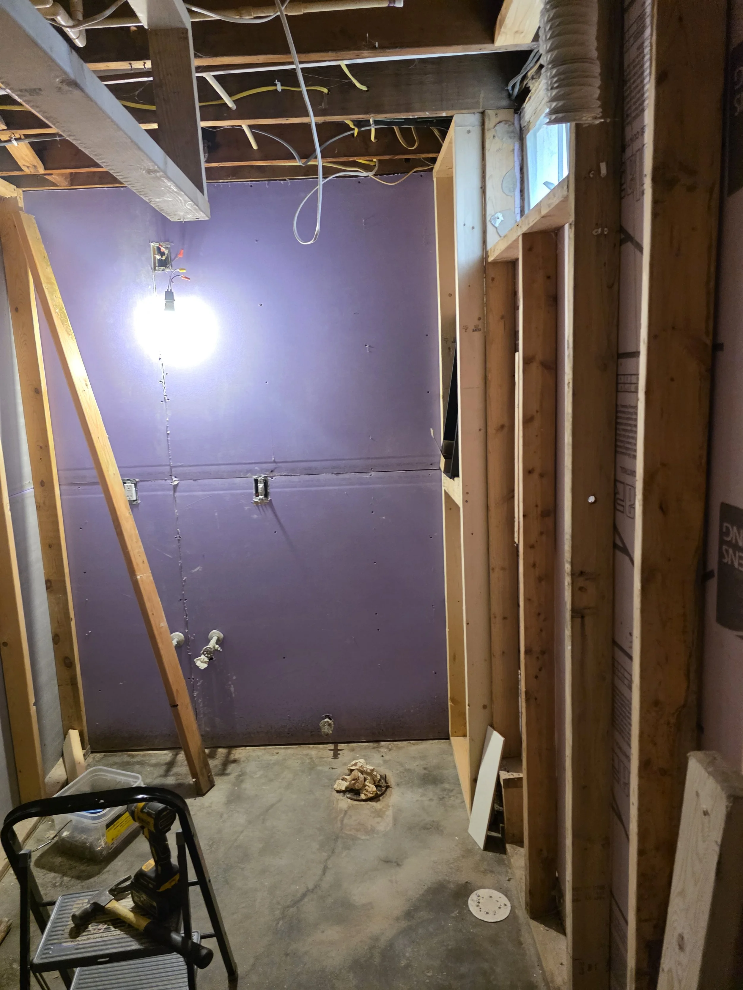 A partially constructed room showing unfinished walls with exposed wooden framing and purple drywall. There are electrical wires hanging from the ceiling and an open window with a curtain. Construction tools and materials are on the floor and a small step ladder is in the foreground.