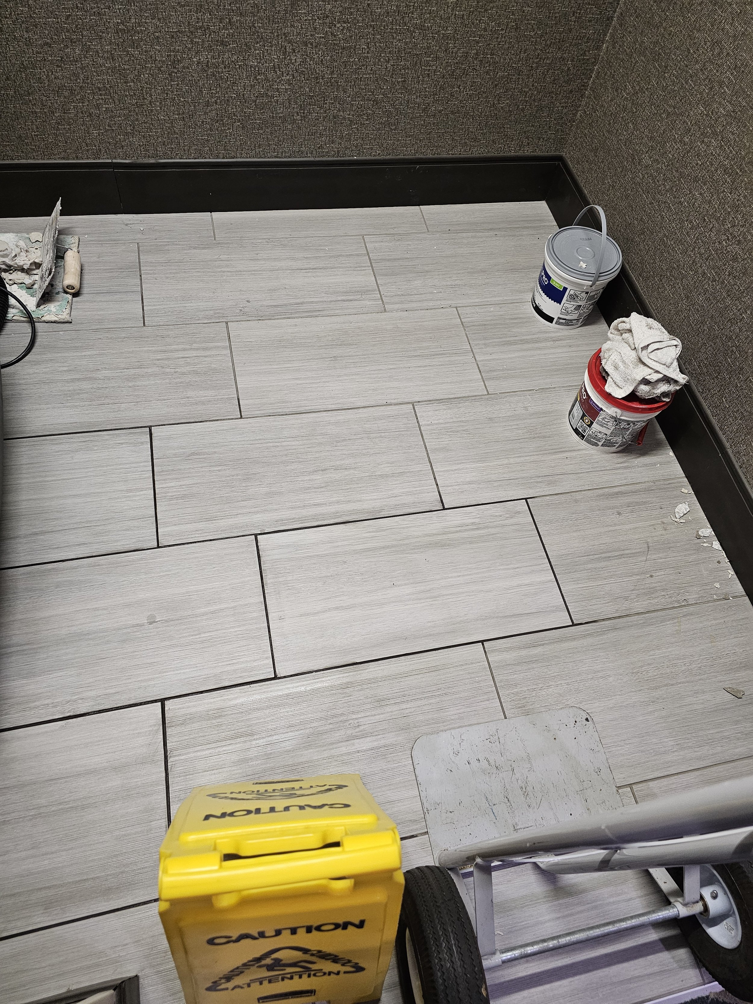 A hospital or medical facility hallway with light-colored tiled floor, dark baseboard, and textured brown walls. Near the right corner, there are two buckets, one white and one red, with cleaning rags on top. A piece of yellow caution tape is in the lower part of the image.