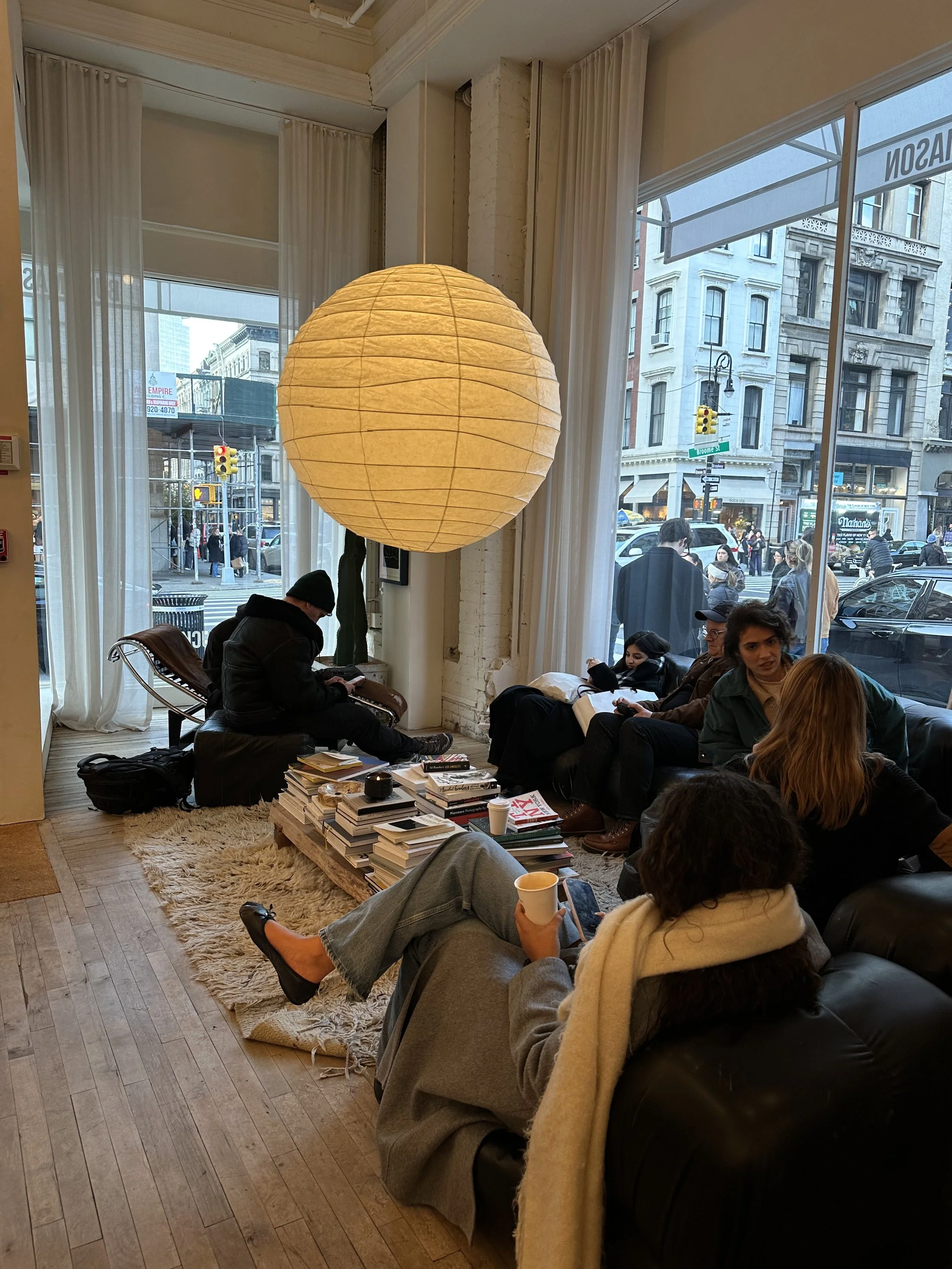 A cozy cafe scene with people sitting on a black leather couch and a person sitting on a chair at a table. There are large windows with white curtains and a warm paper lantern hanging from the ceiling. Outside, pedestrians cross the street and buildings line the sidewalk.
