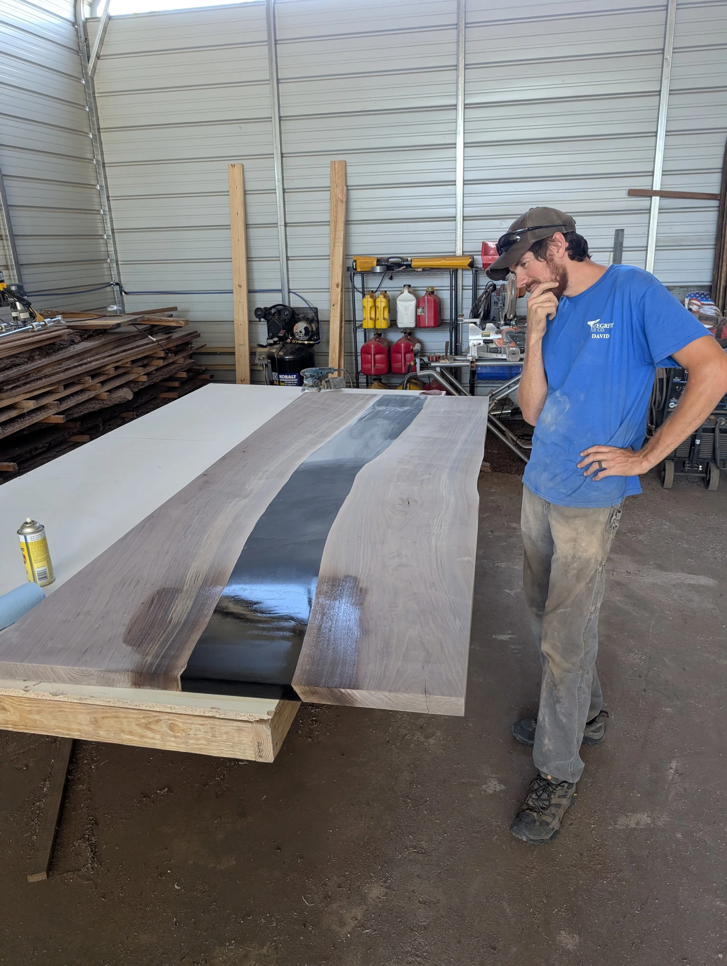 A man wearing a cap, sunglasses, and a blue t-shirt stands in a workshop, looking at a large custom wooden table with a black and gray section, possibly resin or paint, on its surface.
