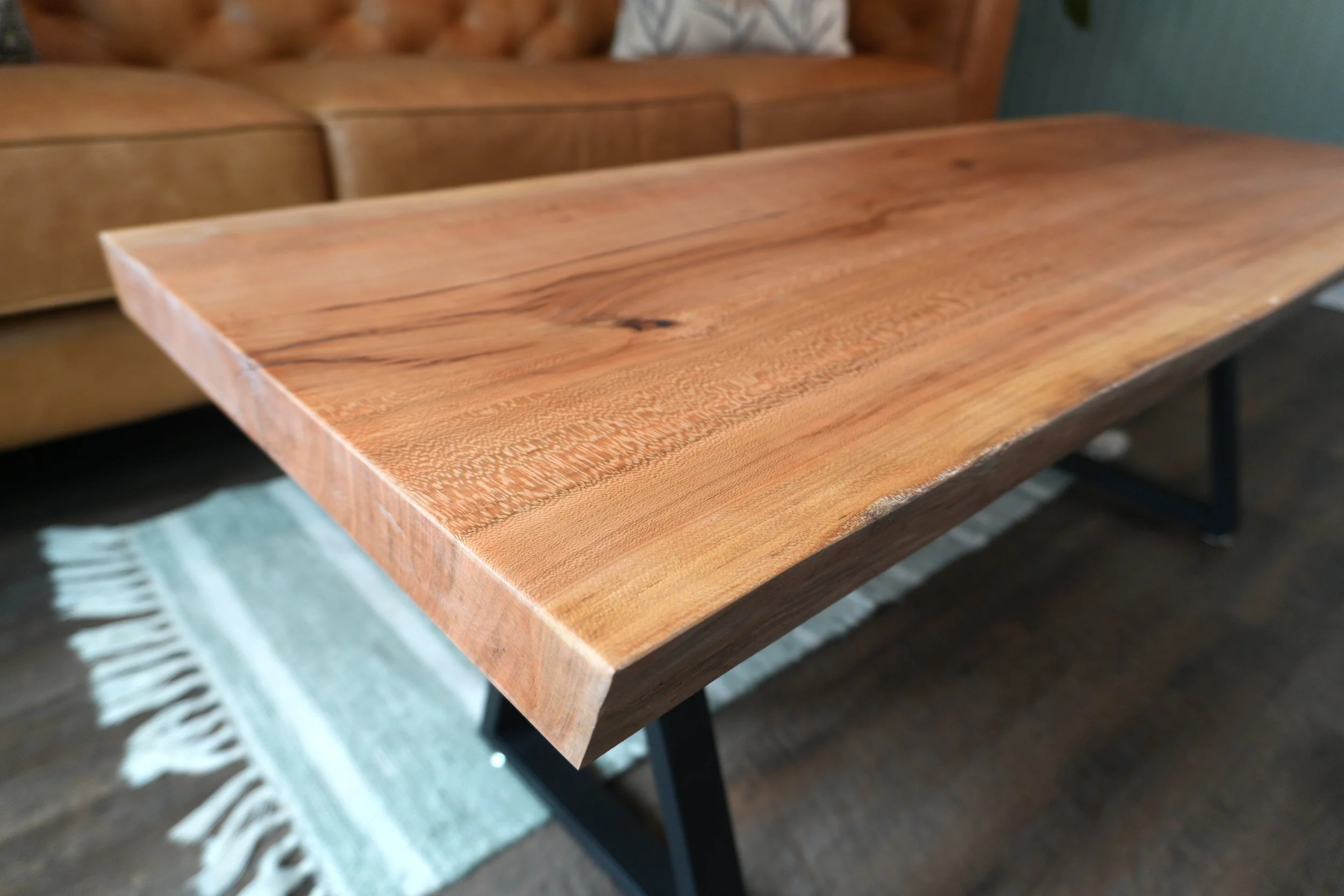 Close-up of a custom wooden table with a natural finish, showing wood grain and knots, with a slightly uneven edge. Part of a patterned throw rug and a dark floor are visible underneath.