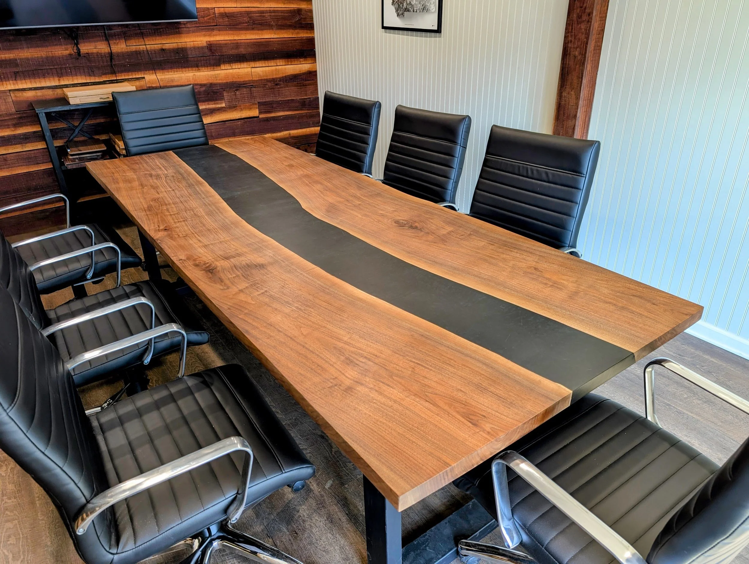 A large wooden conference table with a black runner in the center, surrounded by black leather chairs with metal armrests.