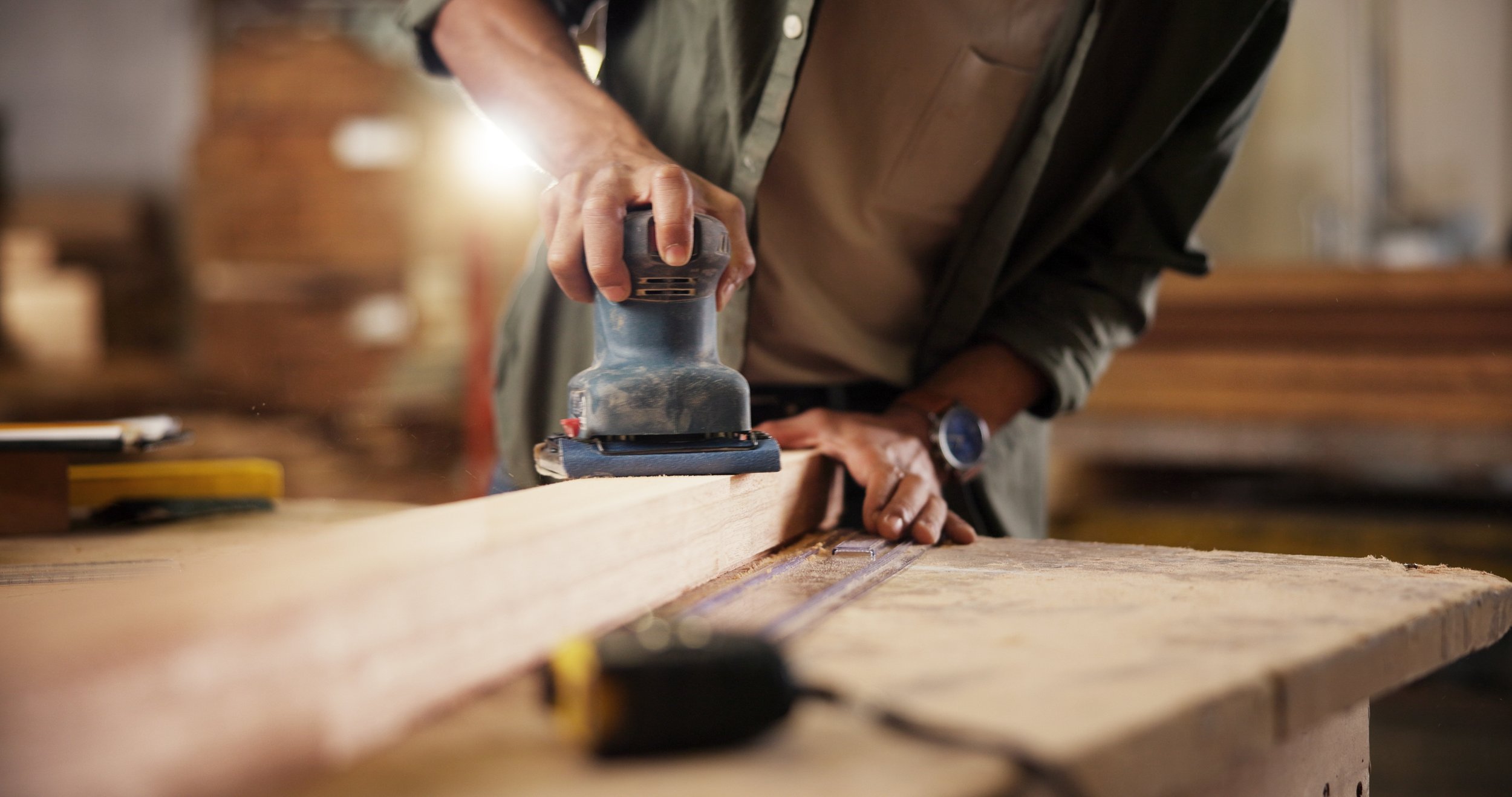 Person sanding a wooden board with a handheld electric sander in a woodworking shop.
