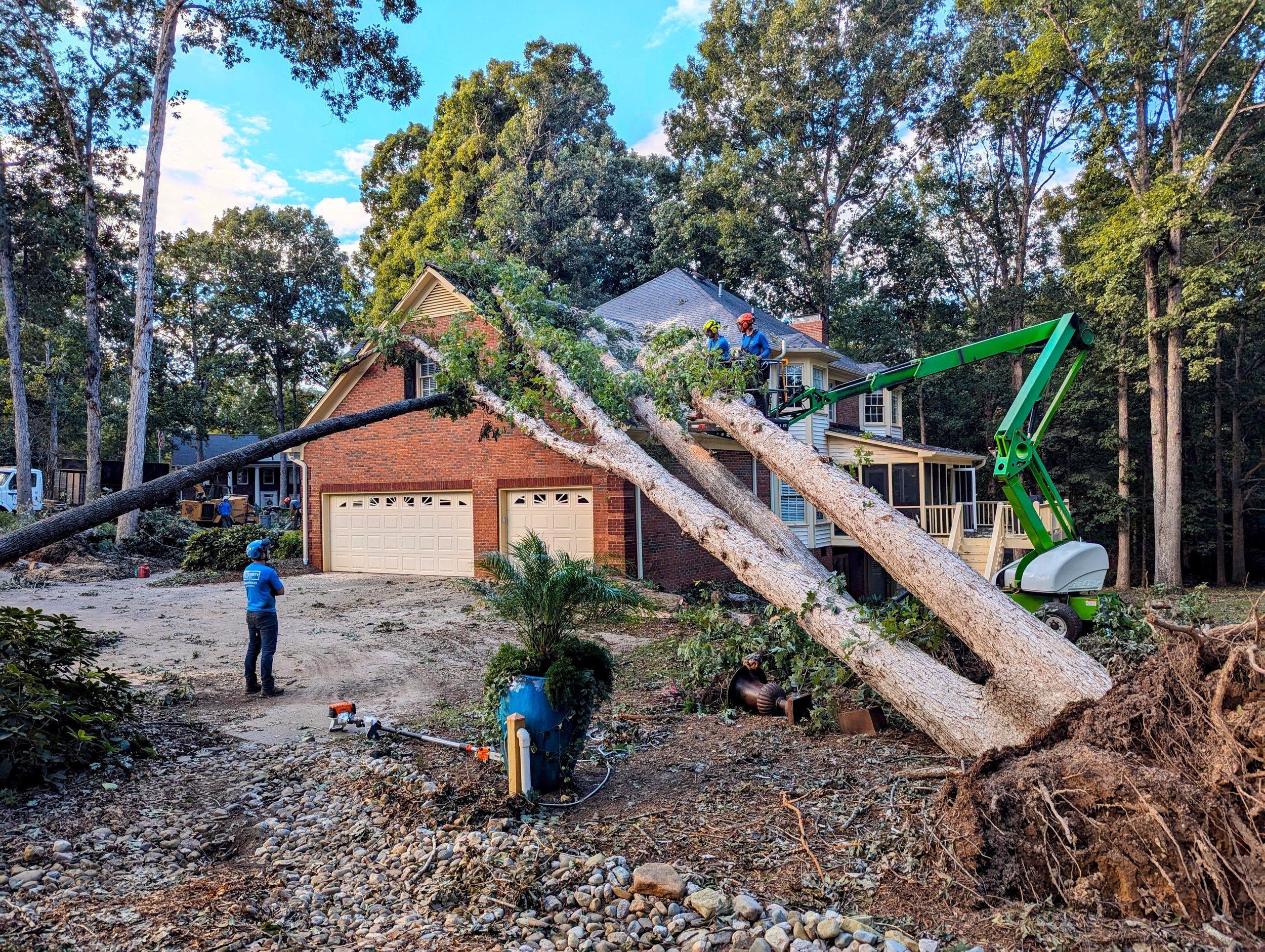 Tree fell on a house, and workers are using a green man lift to remove the branches while other workers are on the ground.