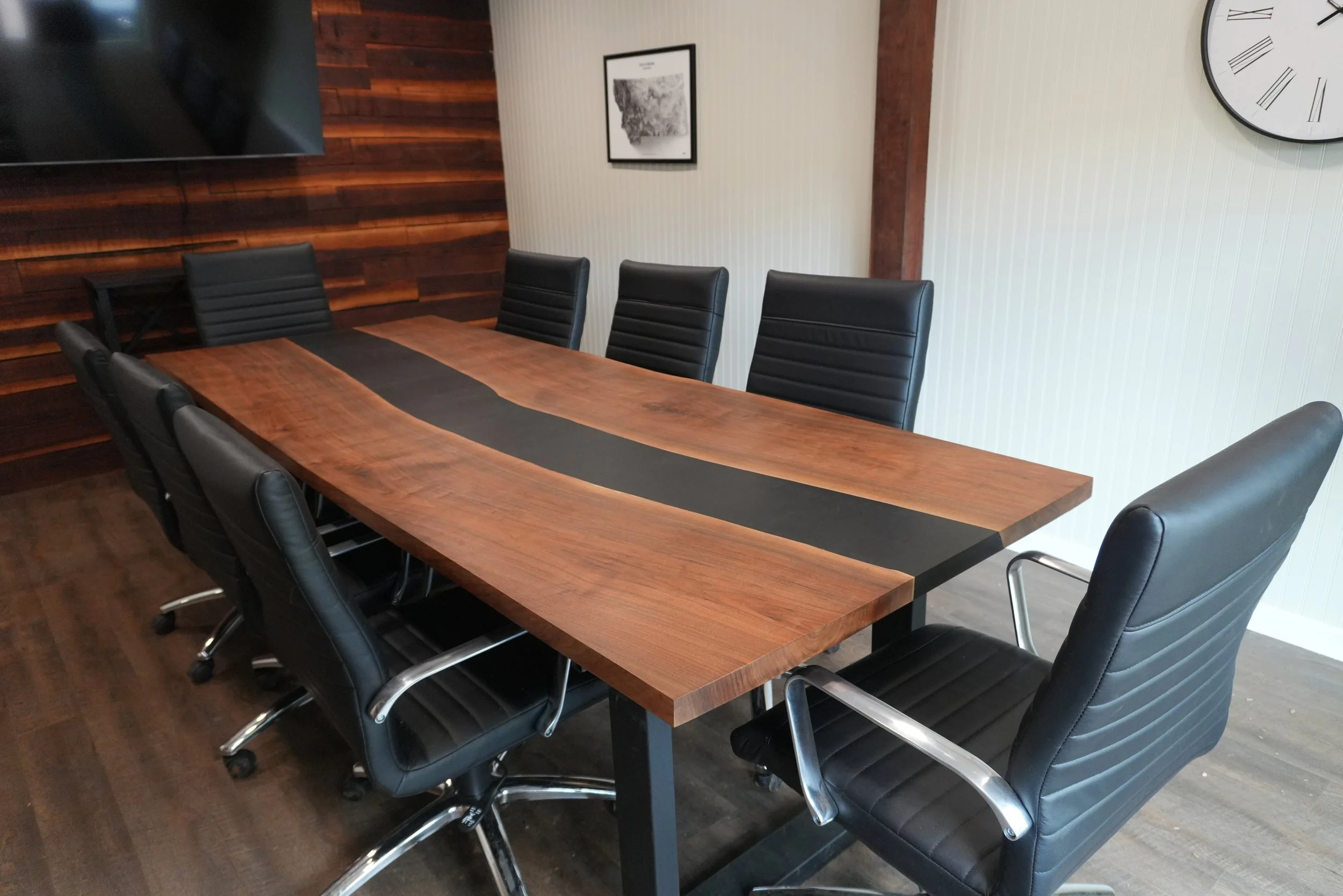 A conference room with a large black walnut wooden table and black leather chairs around it, a wall clock, a framed black-and-white picture, and a wood-paneled wall.