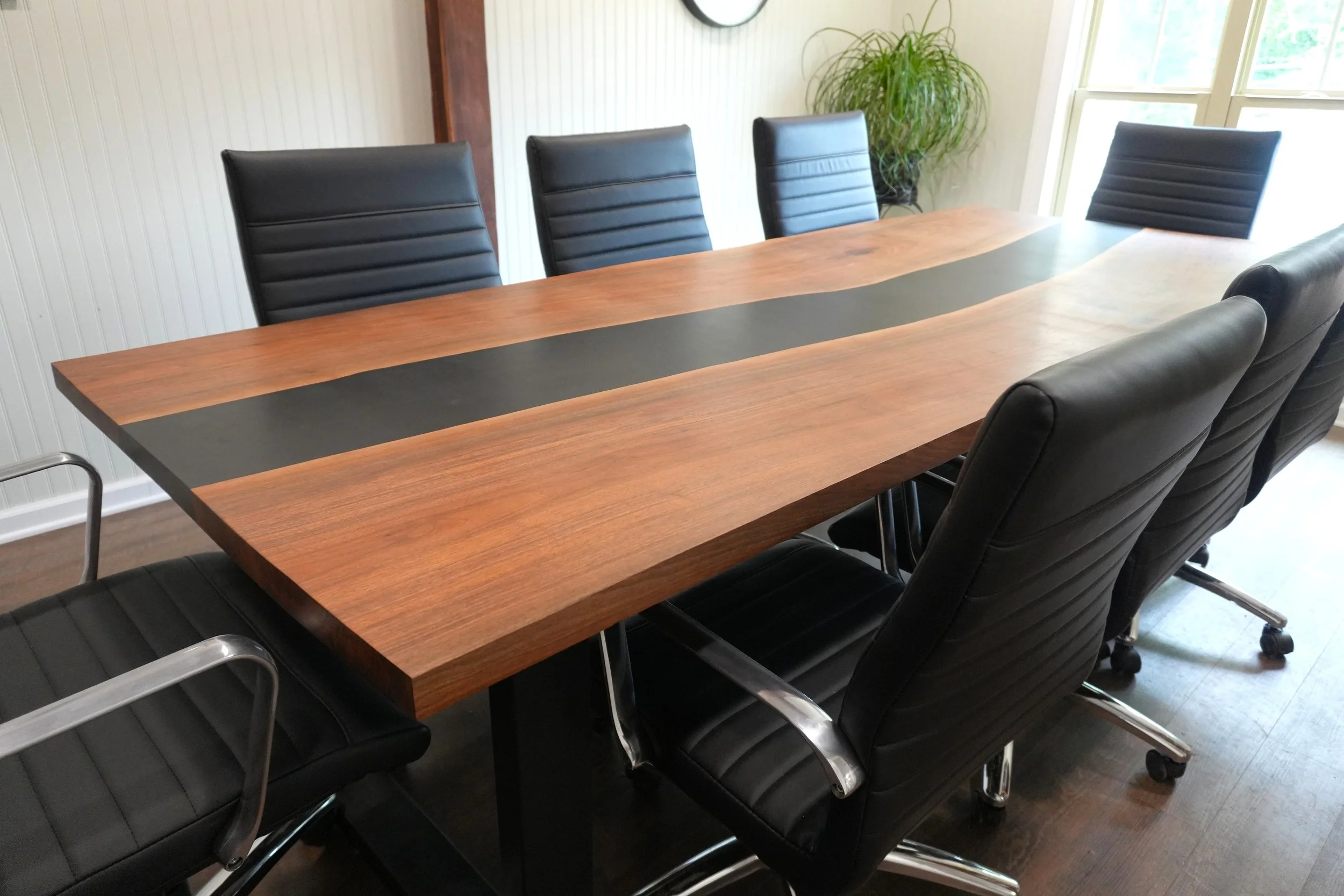 A conference room with a large custom wooden table surrounded by black leather office chairs, a plant in the corner, and windows providing natural light.