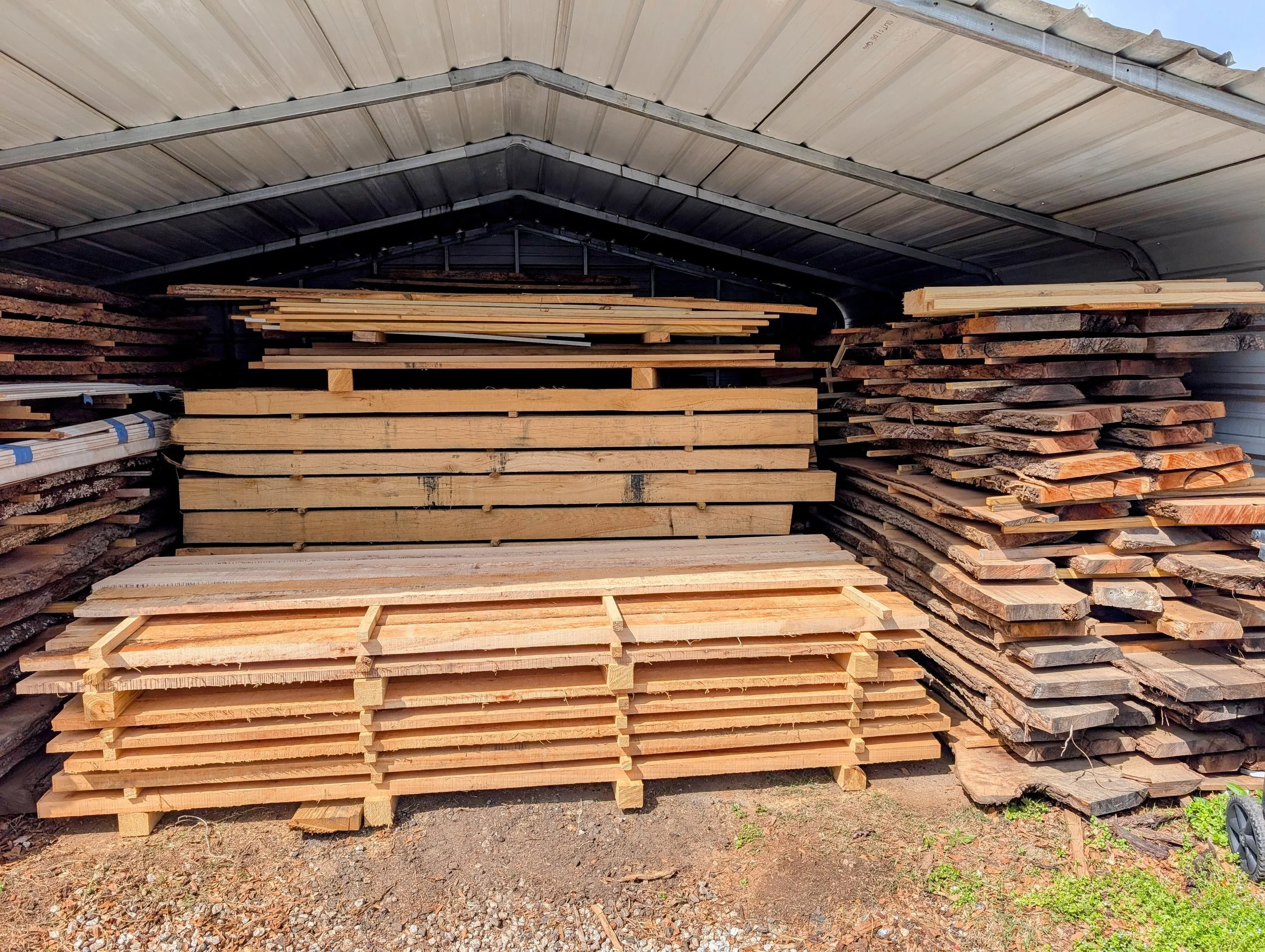 Stacks of locally cut lumber and milled wooden planks stored under a metal roof with an open sky in the background.