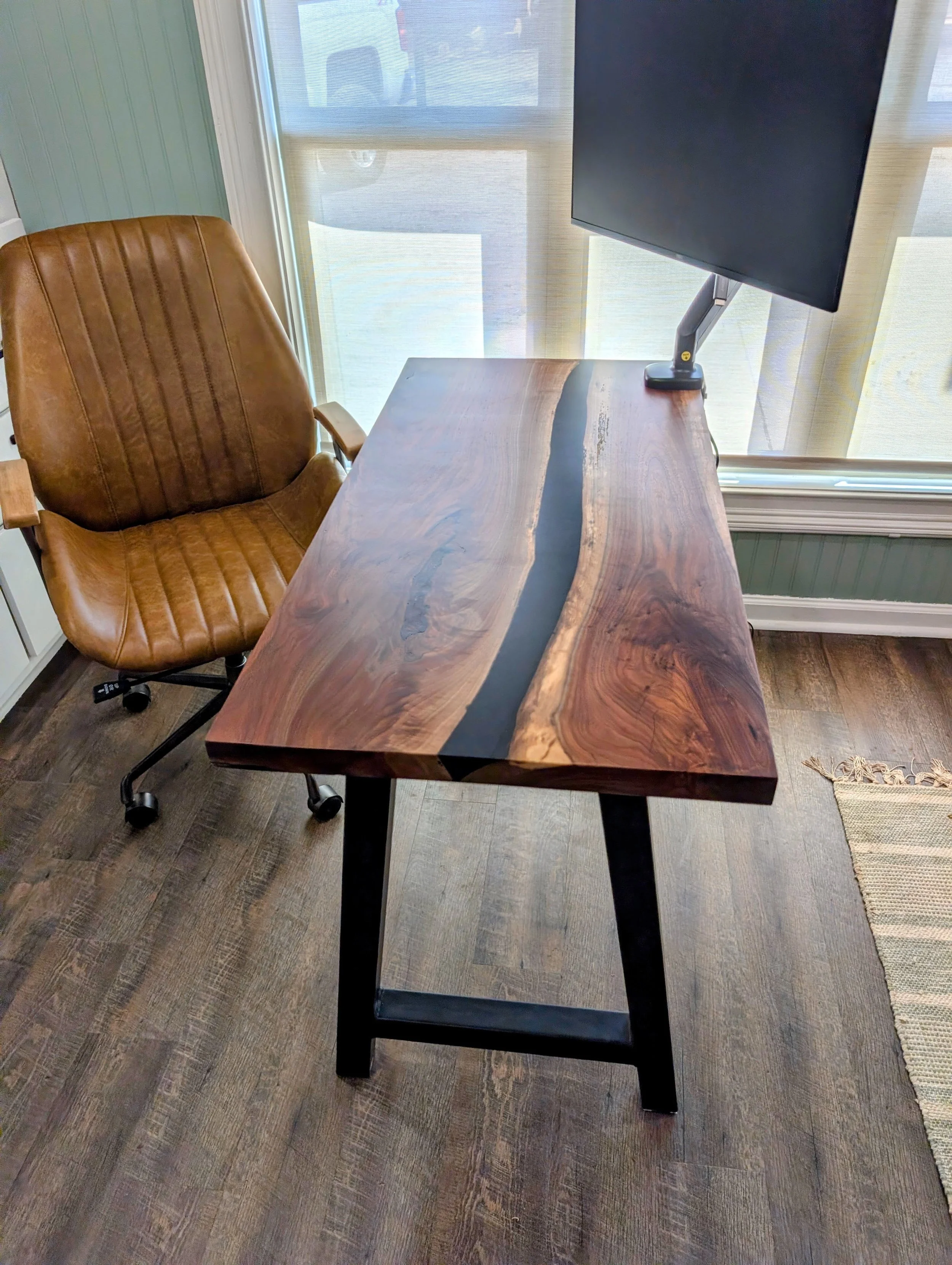 A wooden desk with a black metal frame and a live edge, sitting on a wood-patterned floor. To the left is a brown leather office chair with vertical stitch details. Behind the desk are windows with white blinds, letting in natural light.