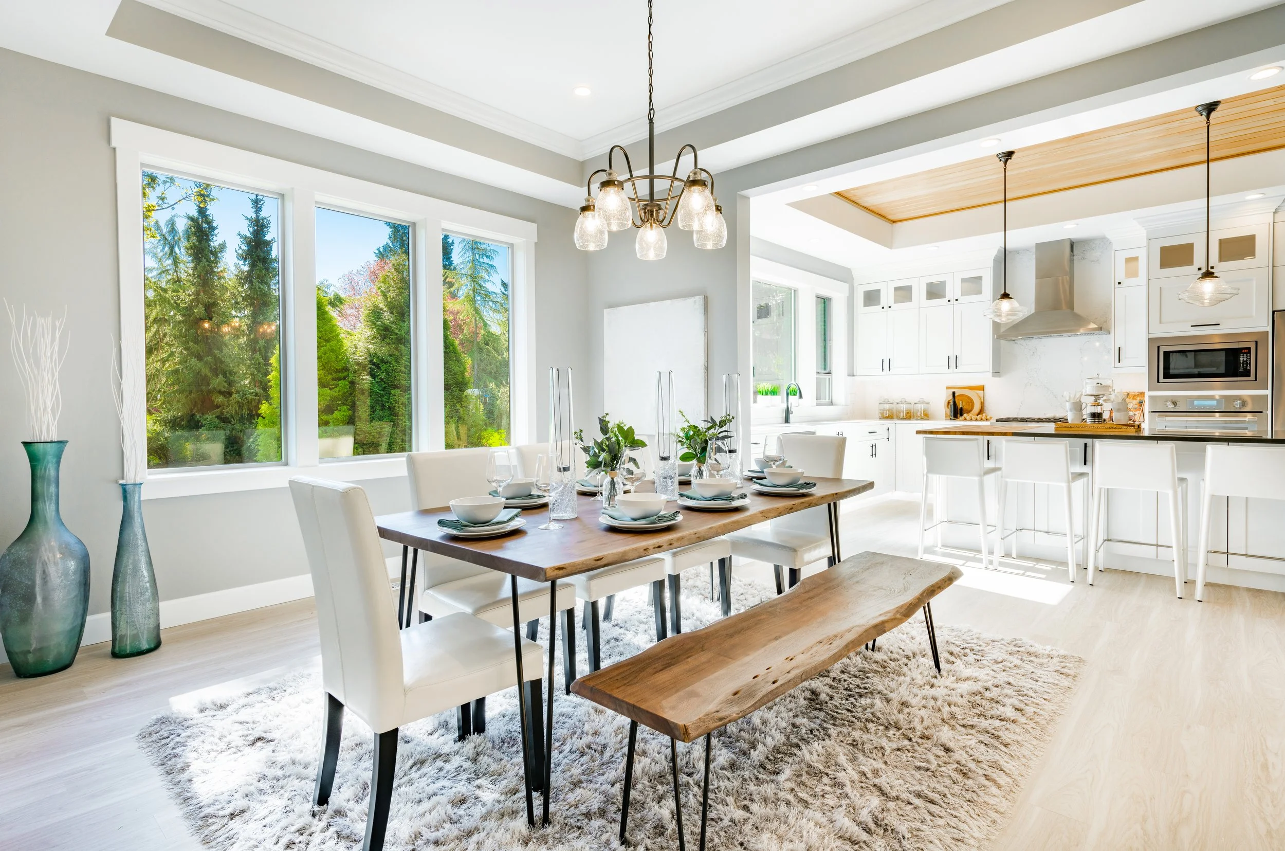 Kitchen and dining room with large windows, white cabinets, wooden accents, and a wood slab dining table with white chairs set for a meal.