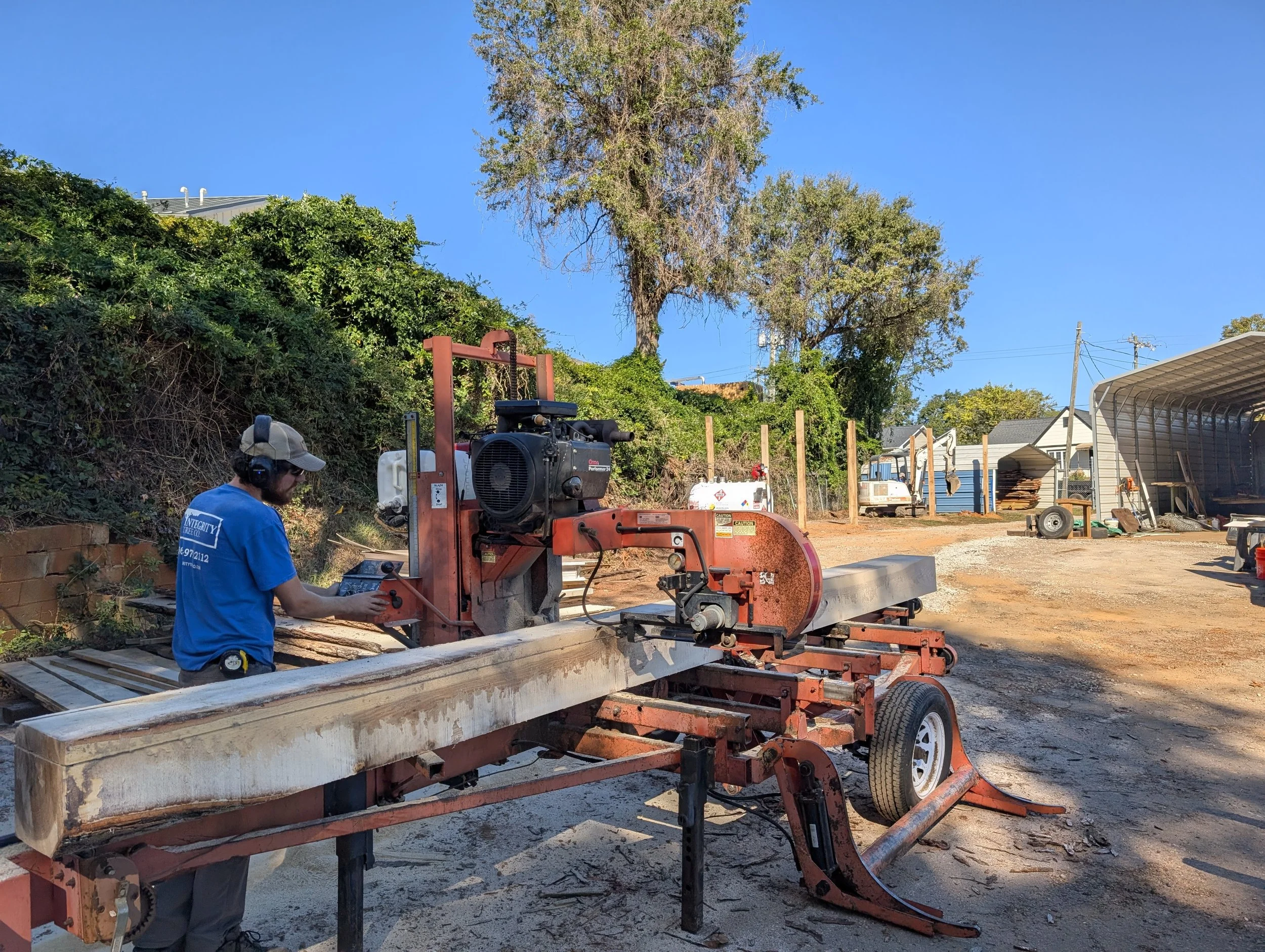 A construction worker operating a wood mill on a worksite with trees and buildings in the background.