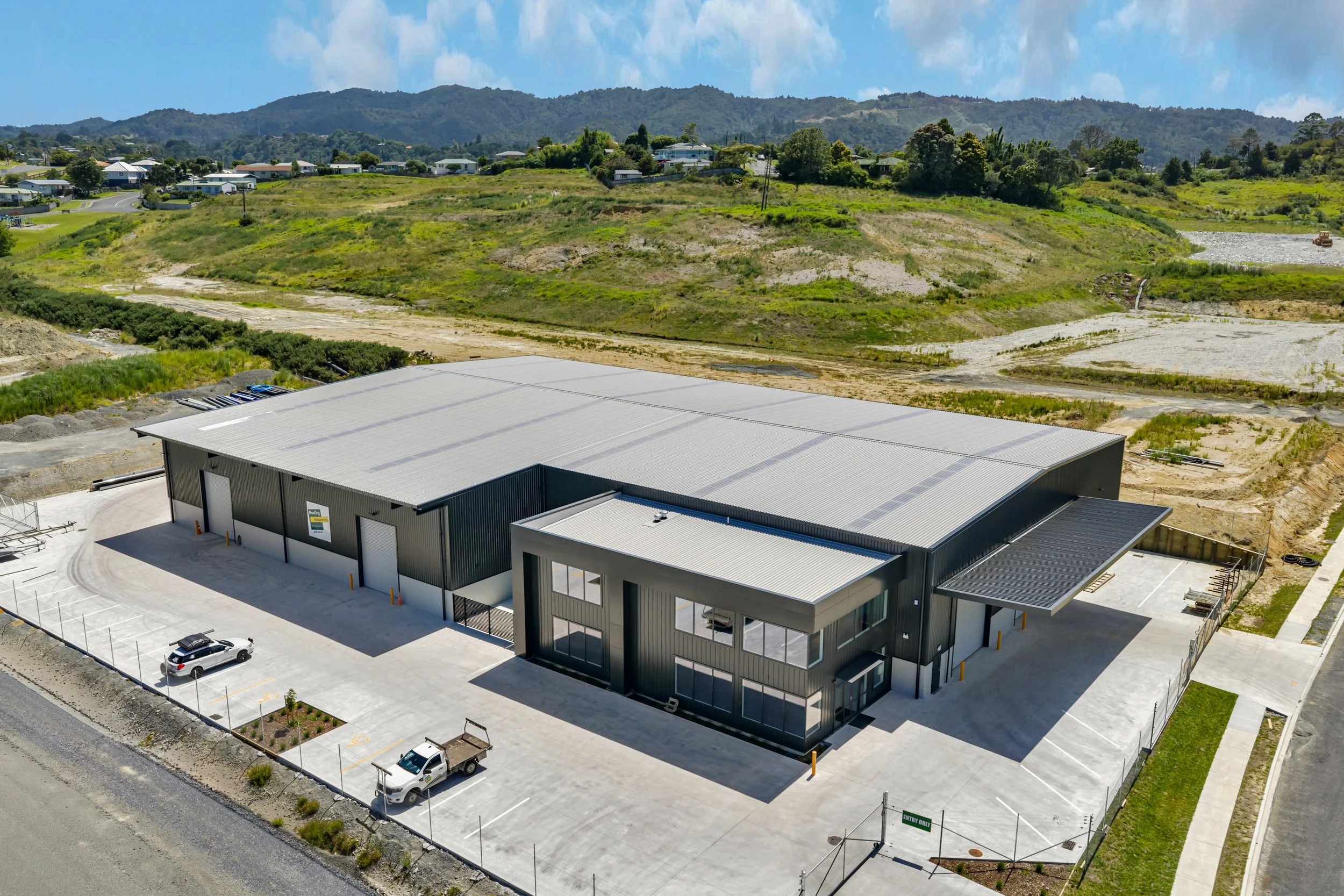 An aerial view of a modern commercial building with a parking lot, set against a green hillside with houses and trees in the background under a partly cloudy sky.