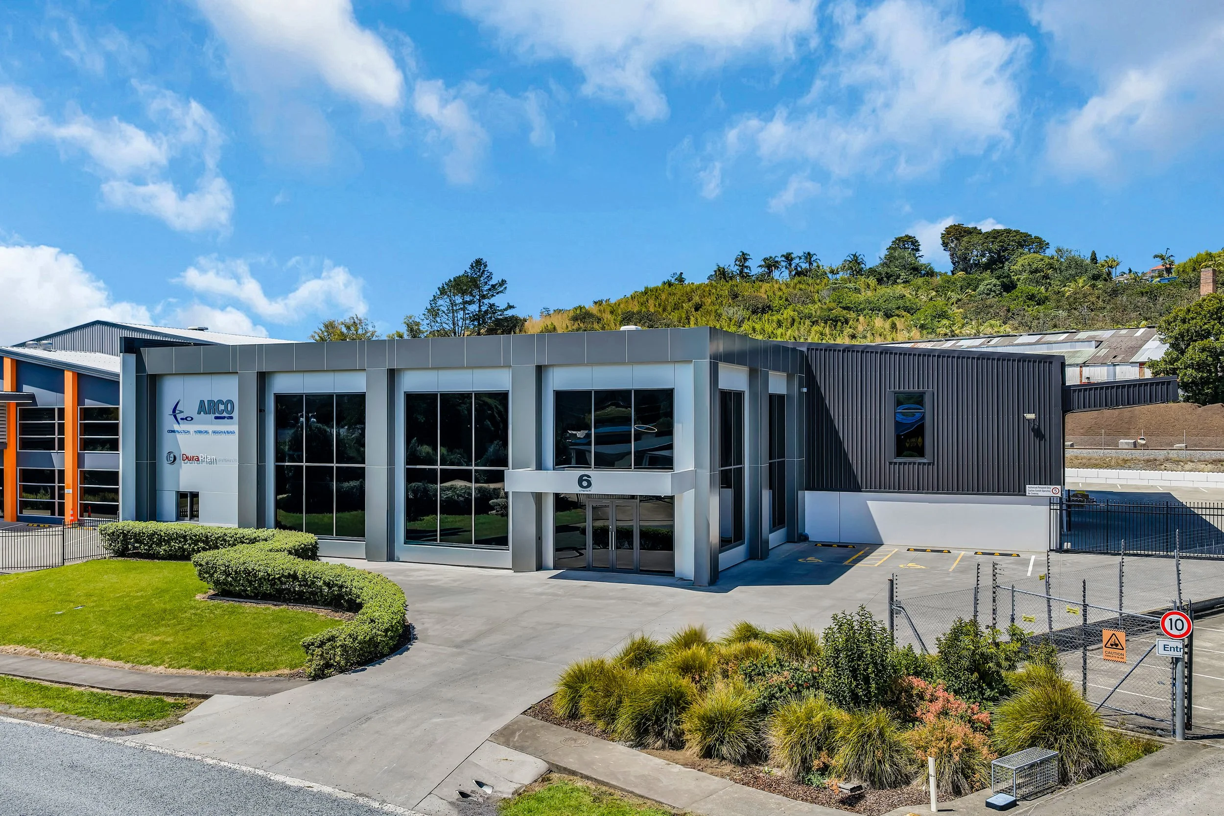 Modern commercial building with large glass windows, situated in a landscaped area with green grass and bushes. The building has signage that reads 'ARCO' and a parking lot with a few marked spaces. The background features a hill with trees and a partly cloudy blue sky.