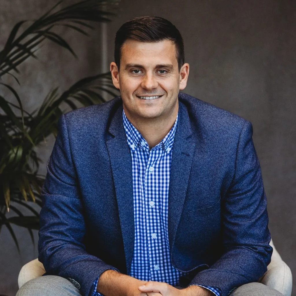 A man with dark hair, wearing a blue blazer and a blue checkered shirt, sitting and smiling in front of a gray background and large green plants.