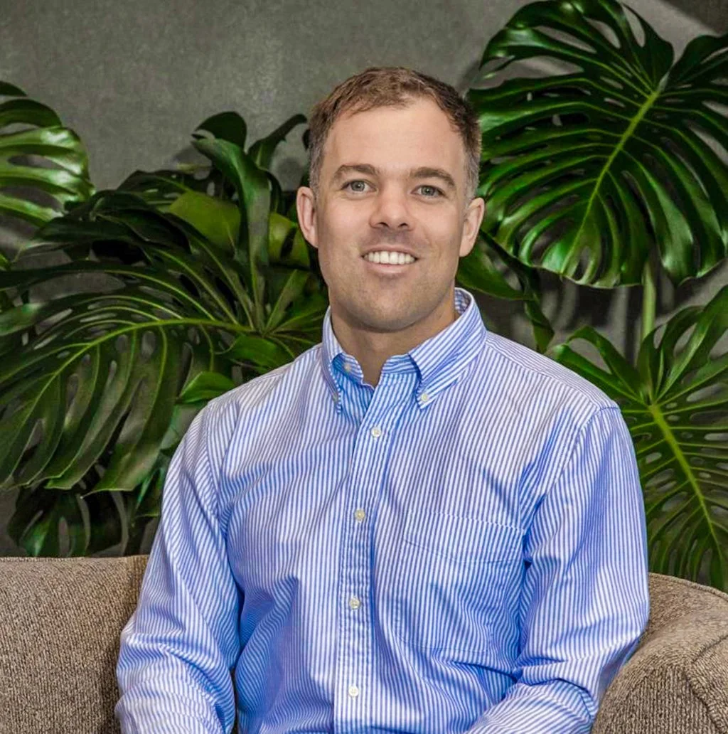A man with short light brown hair and blue eyes, wearing a blue and white striped button-up shirt, sits on a beige couch in front of large green tropical plants.