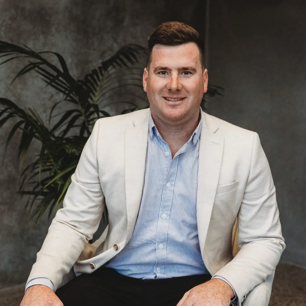 A man wearing a white blazer and blue striped shirt sitting in front of a green plant and a gray textured wall.