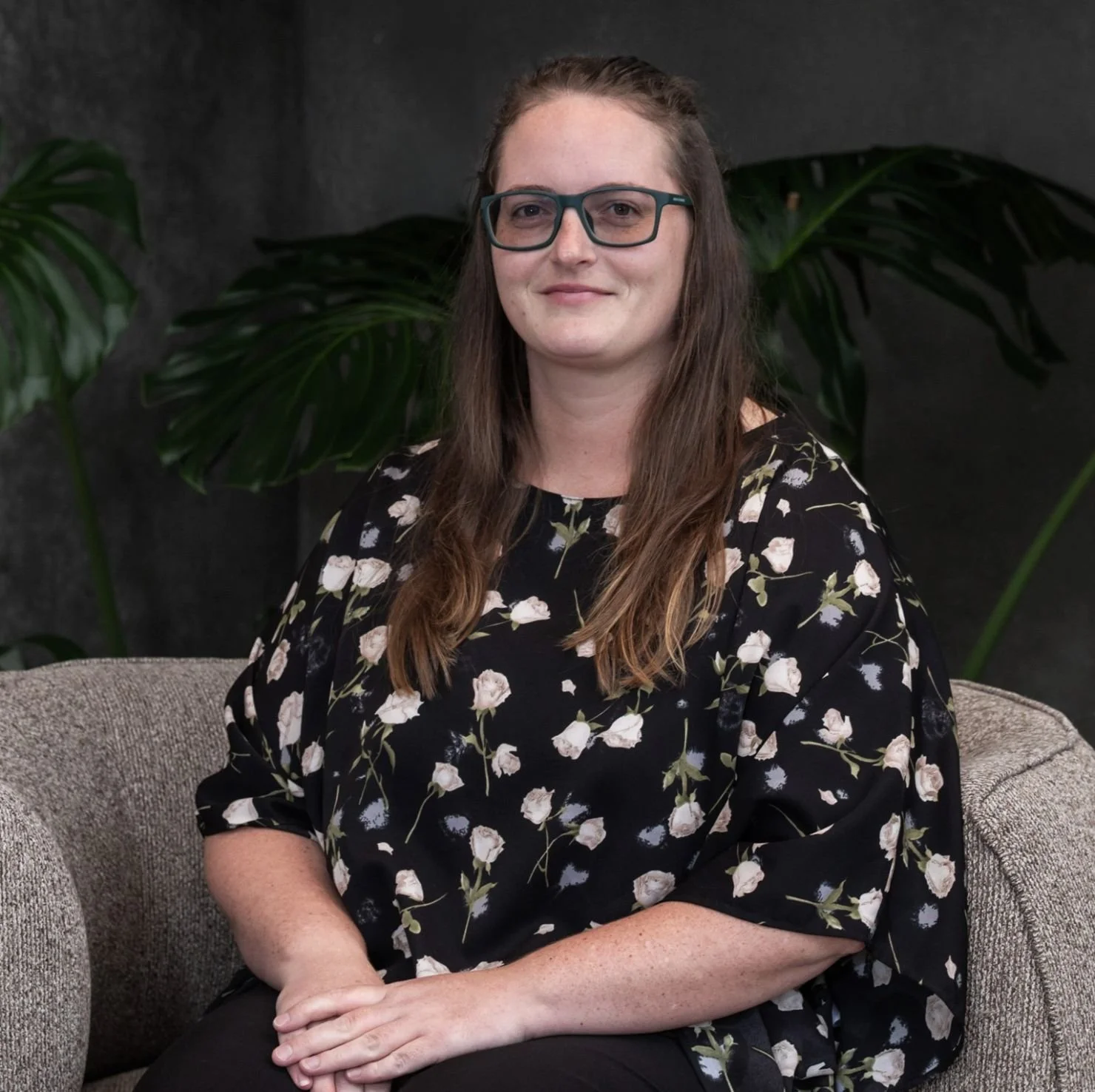A woman with long brown hair, glasses, wearing a black floral top, sitting on a beige couch with large green leaves in the background.