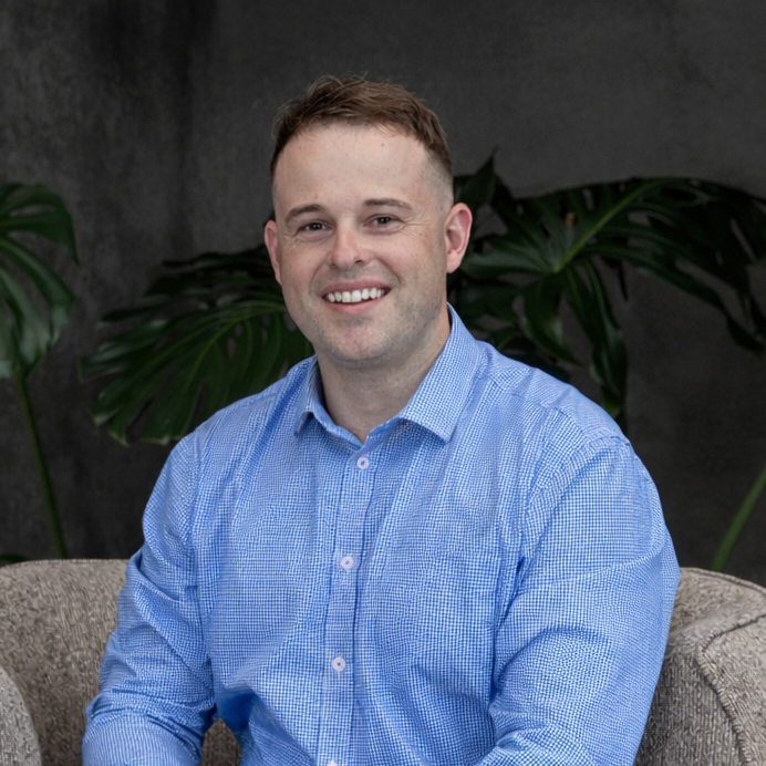 Portrait of a young man with short, light brown hair, wearing a blue checkered dress shirt, sitting on a beige couch with large green plants in the background.