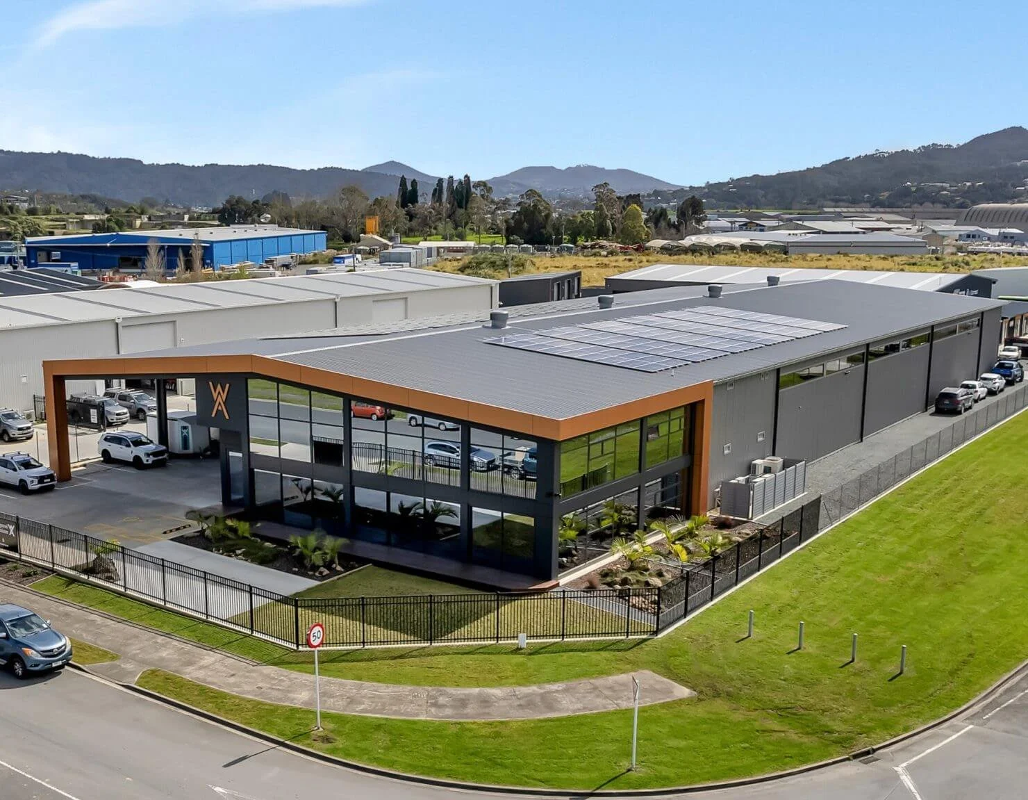 A modern industrial building with solar panels on the roof, a fenced perimeter, surrounded by green grass, parking lot, and mountains in the background.