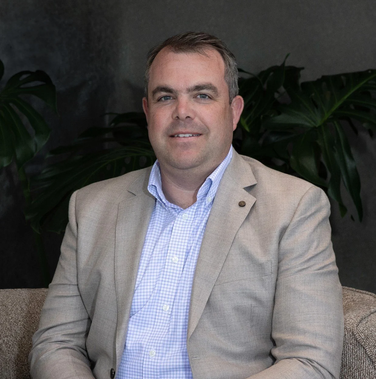 A man wearing a beige blazer and light blue checkered shirt sitting in front of green plants and a dark background.