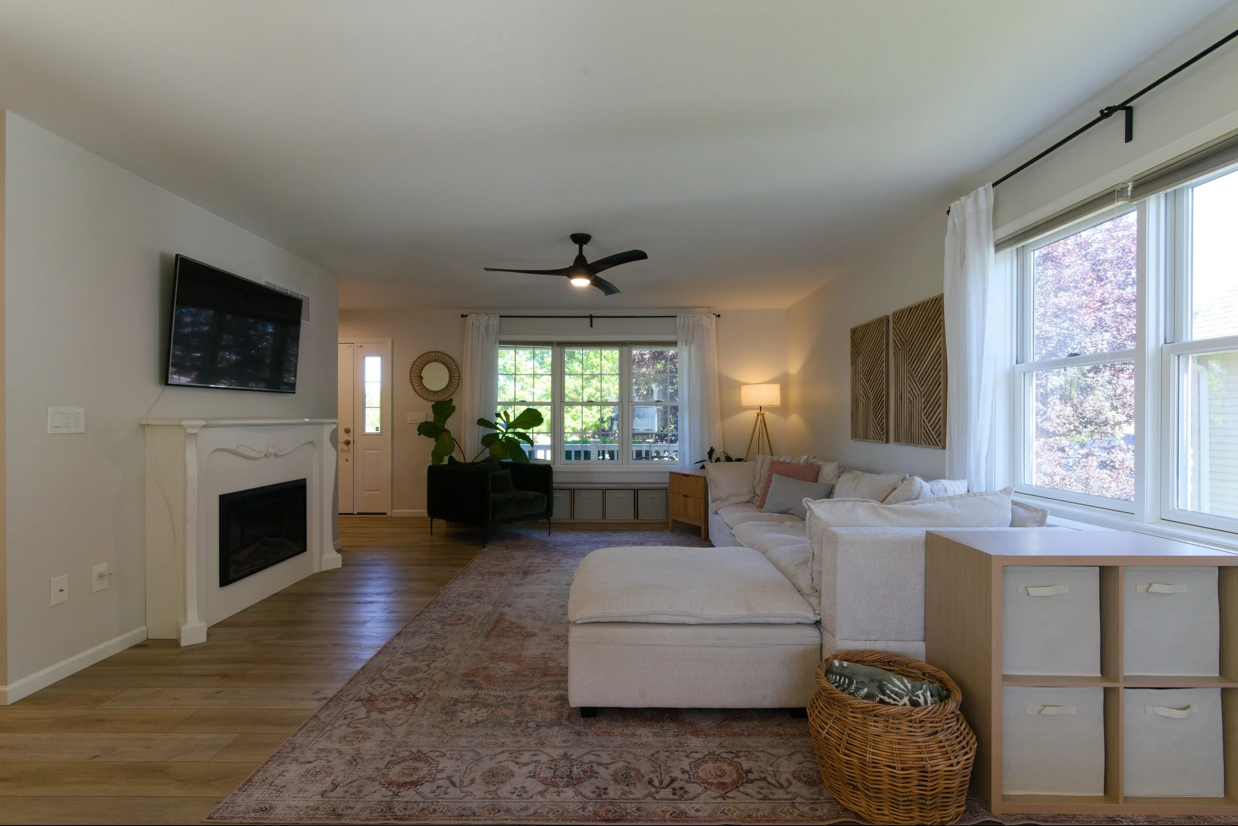 Living room with large windows, white curtains, beige sofa, black armchair, wooden side table, television mounted on wall, ceiling fan, lamp, potted plant, decorative wall art, and woven basket on floor.