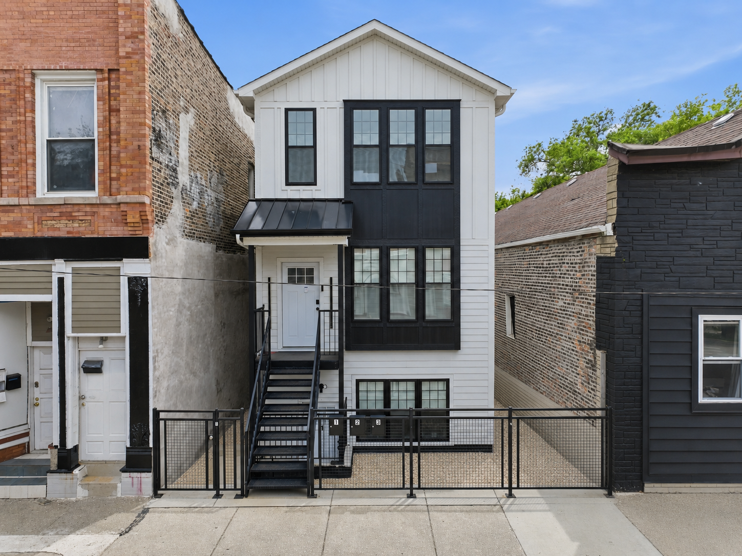 A modern three-story house with white siding and black window frames, located between older brick and dark gray houses, with an external black staircase leading to the second floor, and a black metal fence in front.