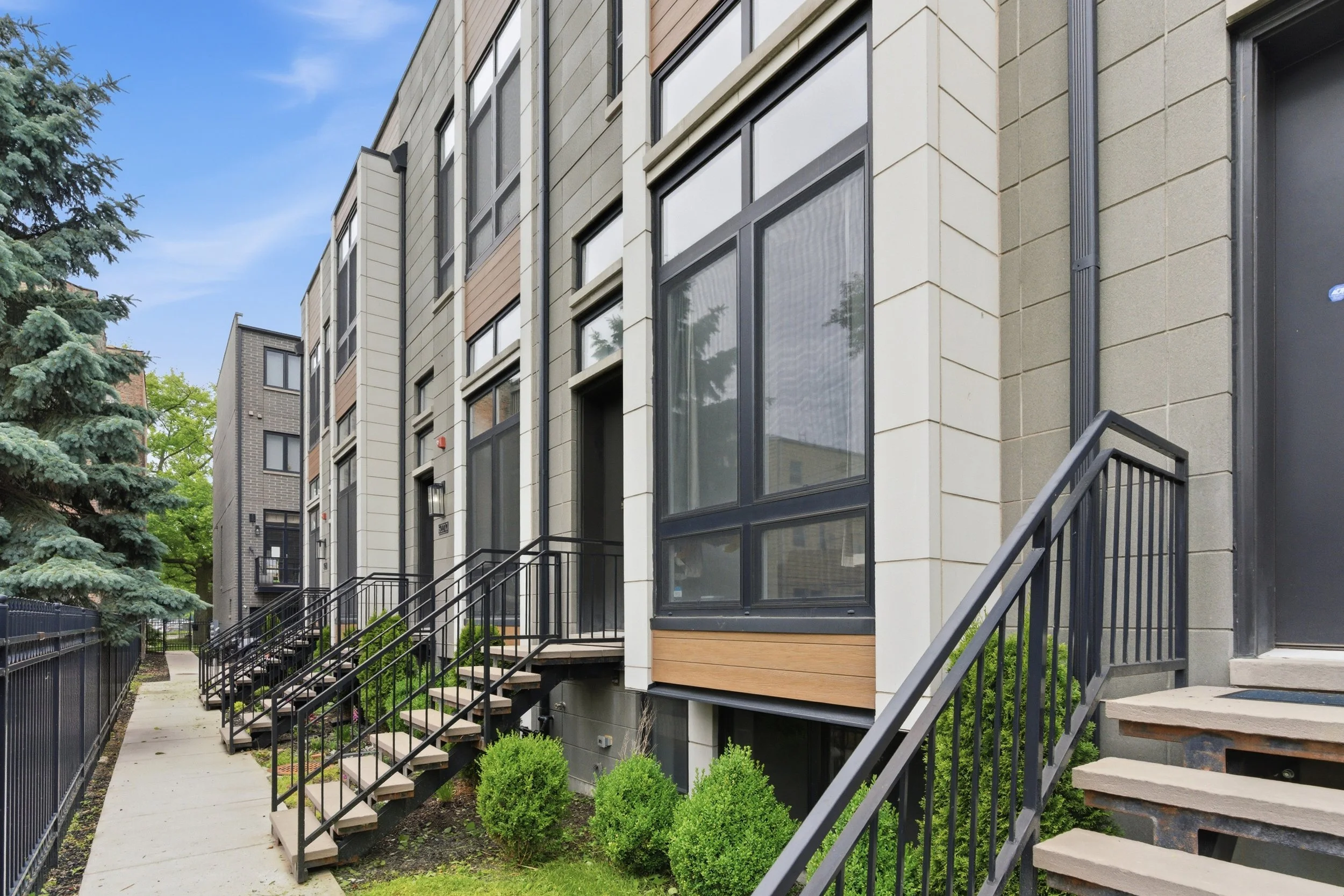 Exterior view of modern multi-story residential buildings with stairs and greenery in the foreground, and a blue sky with clouds in the background.