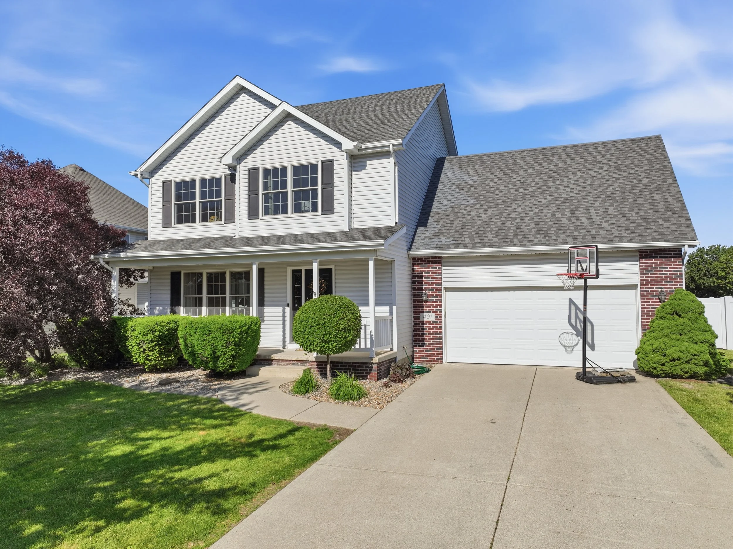A two-story house with white siding, black shutters, and a gable roof, with a basketball hoop on the driveway, a small front porch, green bushes, and trees nearby under a blue sky.