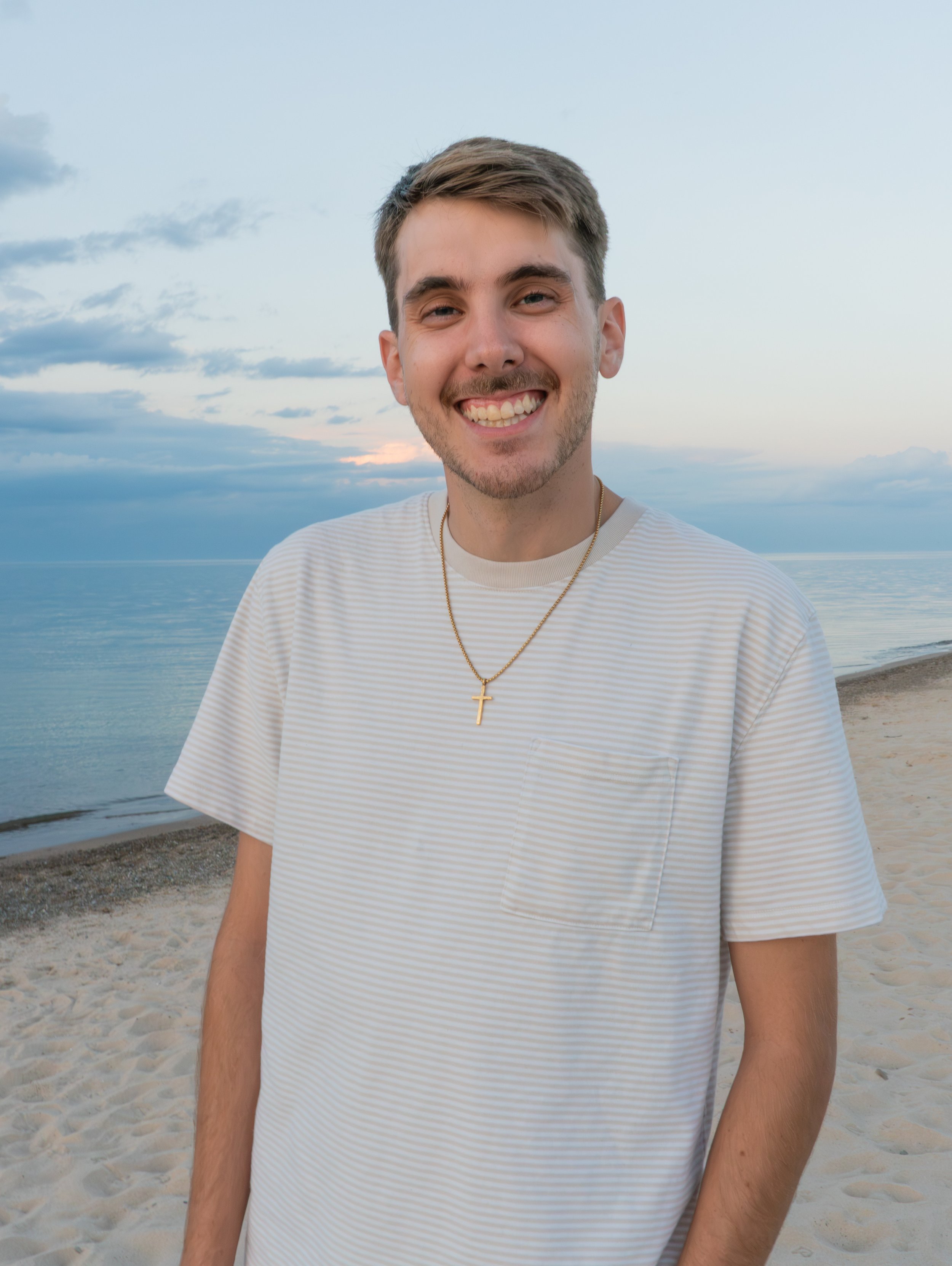 A young man with a smile, wearing a striped white t-shirt and a gold cross necklace, standing on a sandy beach at sunset with a body of water and cloudy sky in the background.