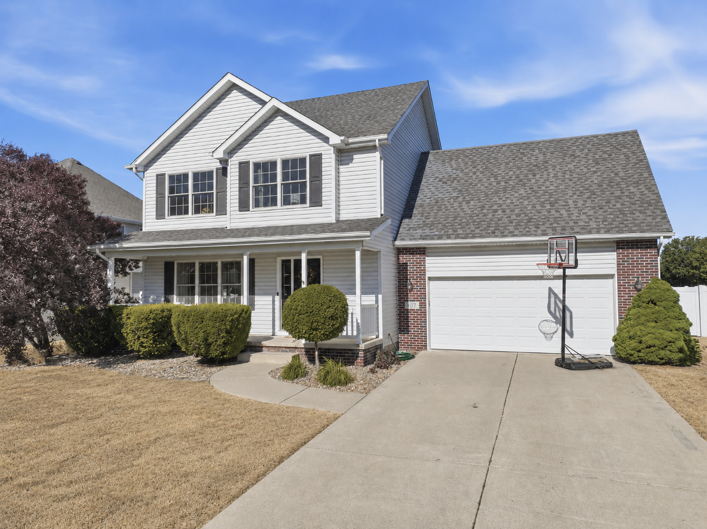 Two-story suburban house with white siding, gray roof, attached garage with a basketball hoop in driveway, front yard with trimmed bushes and a tree, under a blue sky.