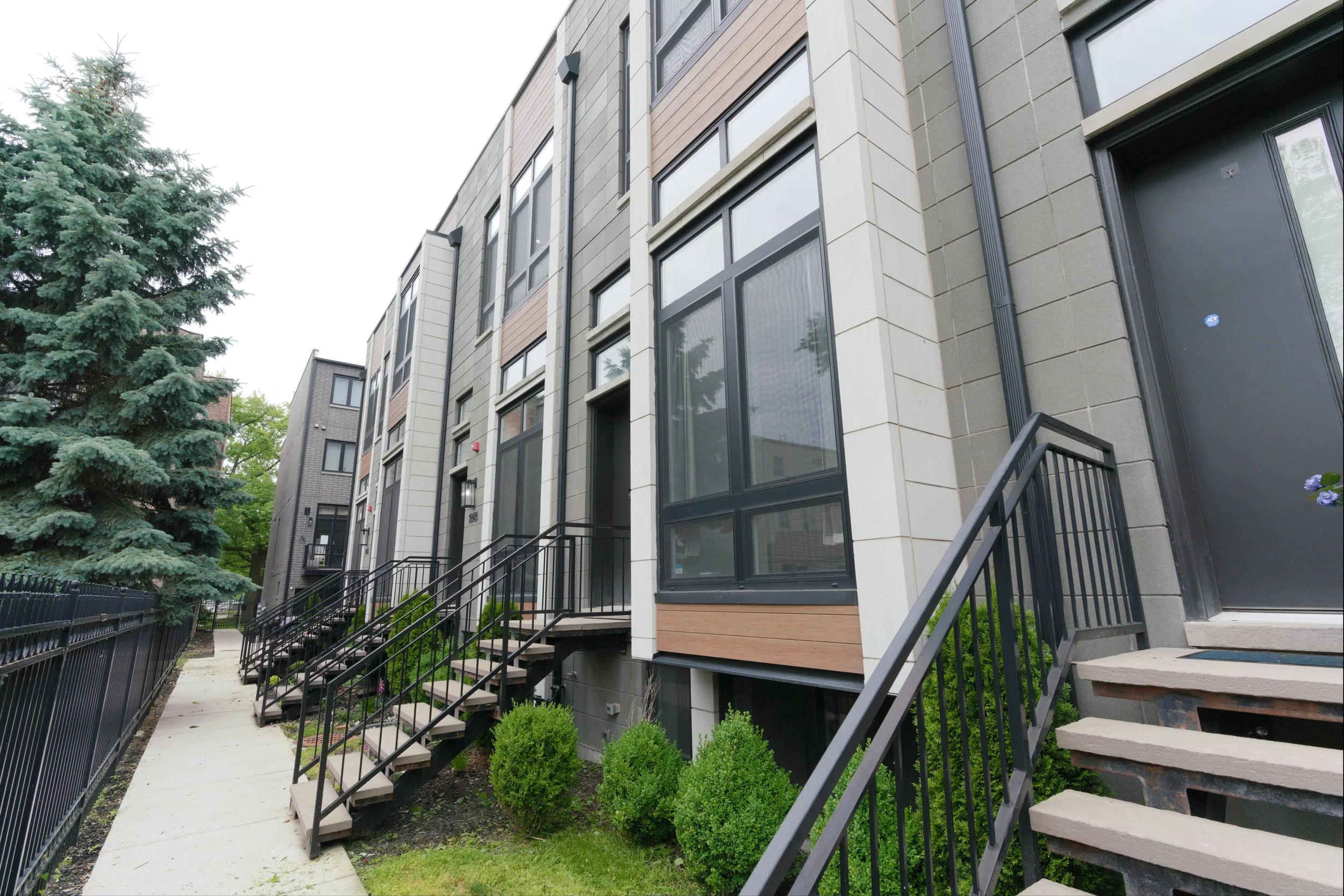 Exterior view of modern multi-story apartment buildings with large windows and stairs leading to entrances, surrounded by green bushes and a large evergreen tree.