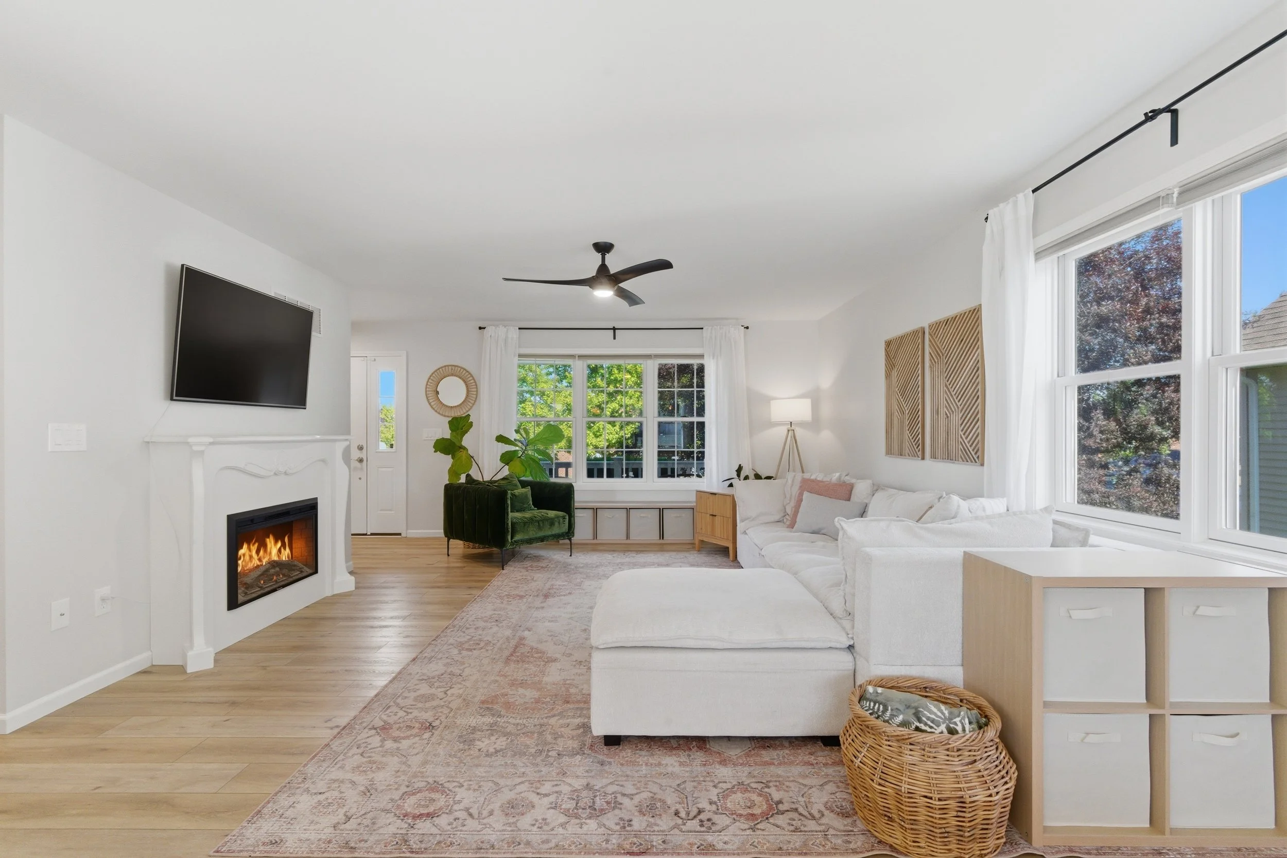 Bright living room with large windows, white walls, a fireplace, a flat-screen TV mounted on the wall, a green velvet armchair, a white sofa, a standing lamp, and a woven basket in front of a side table.