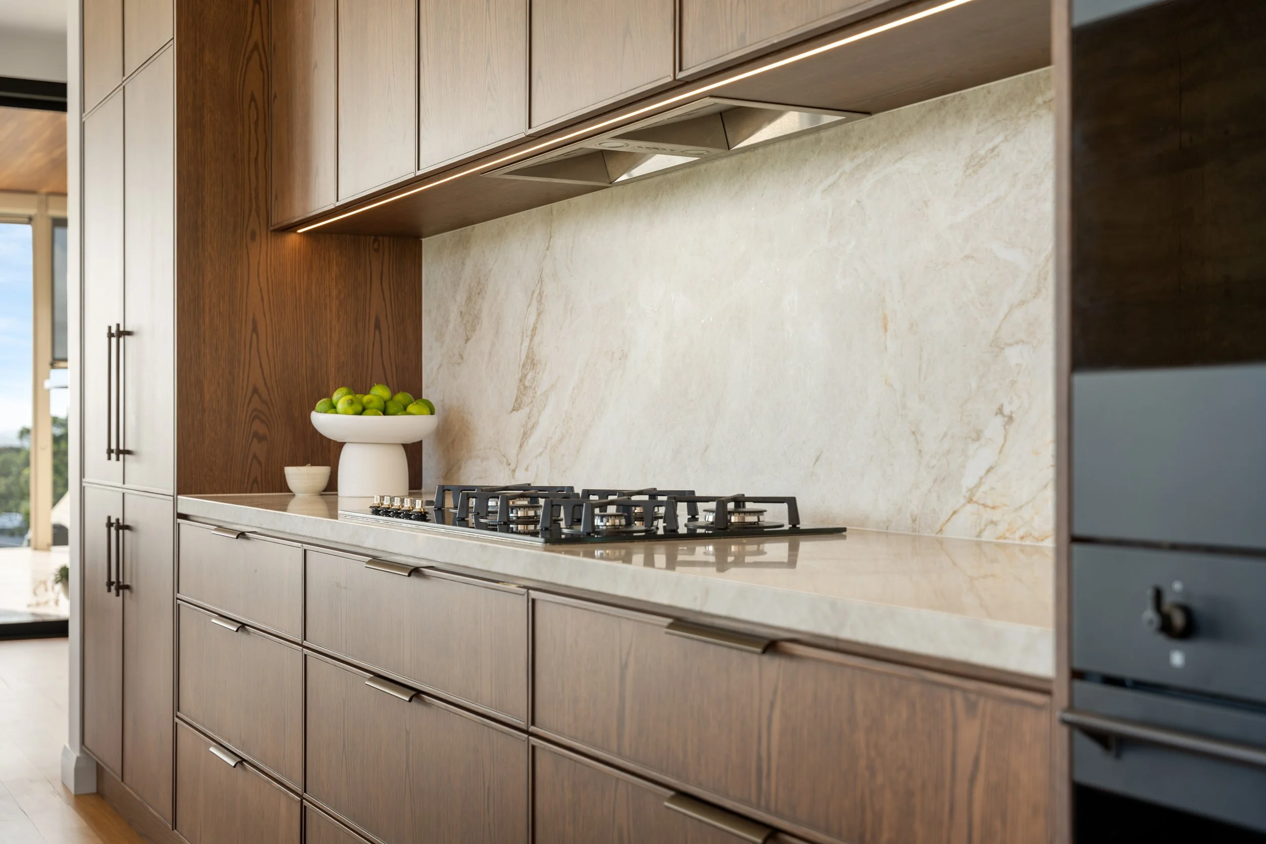 Modern kitchen with wooden cabinets, a beige marble backsplash, a stovetop, and a bowl of green apples.