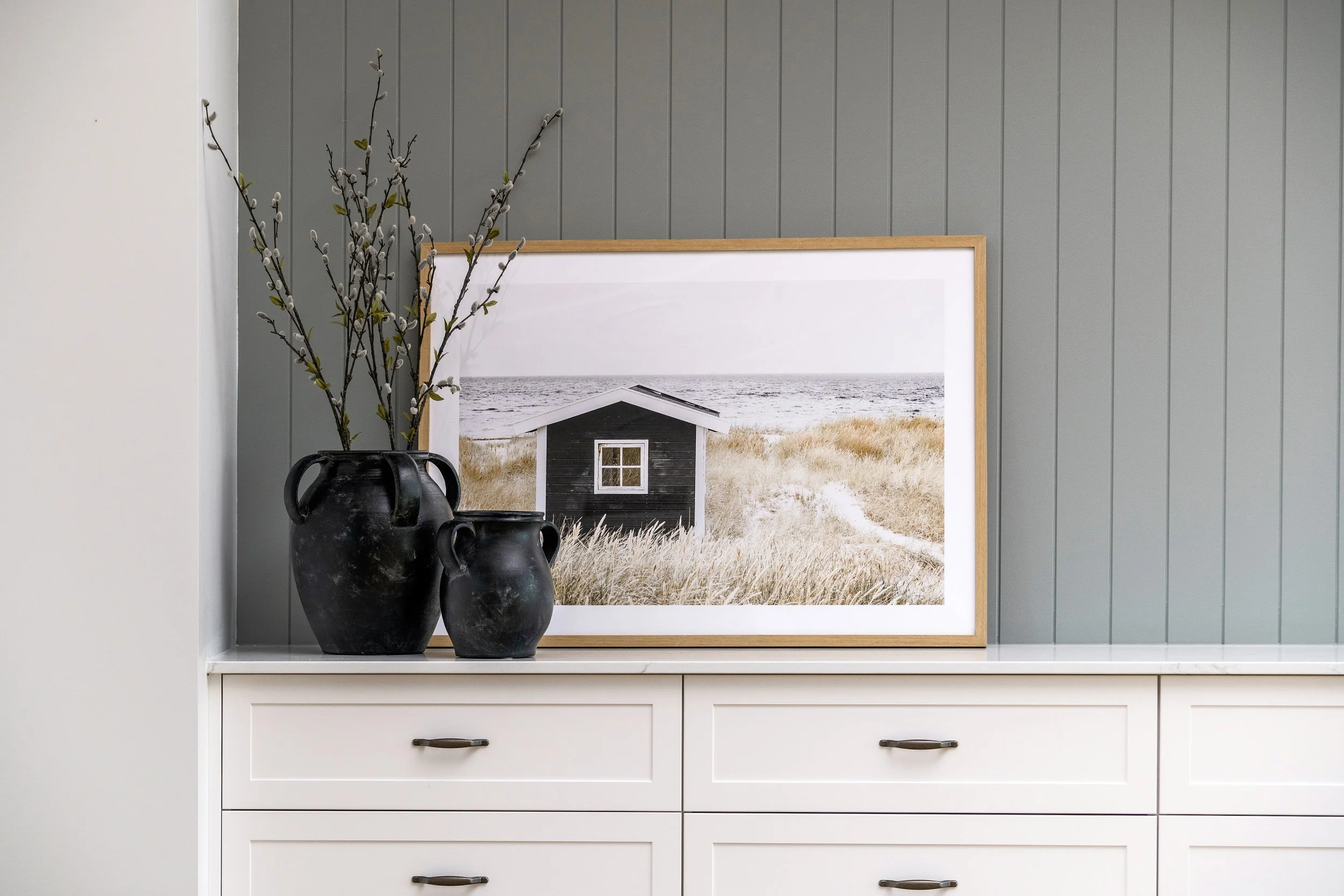 Vase with flowering branches and framed landscape photograph of a small house near the ocean, all positioned on a white dresser against a gray paneled wall.