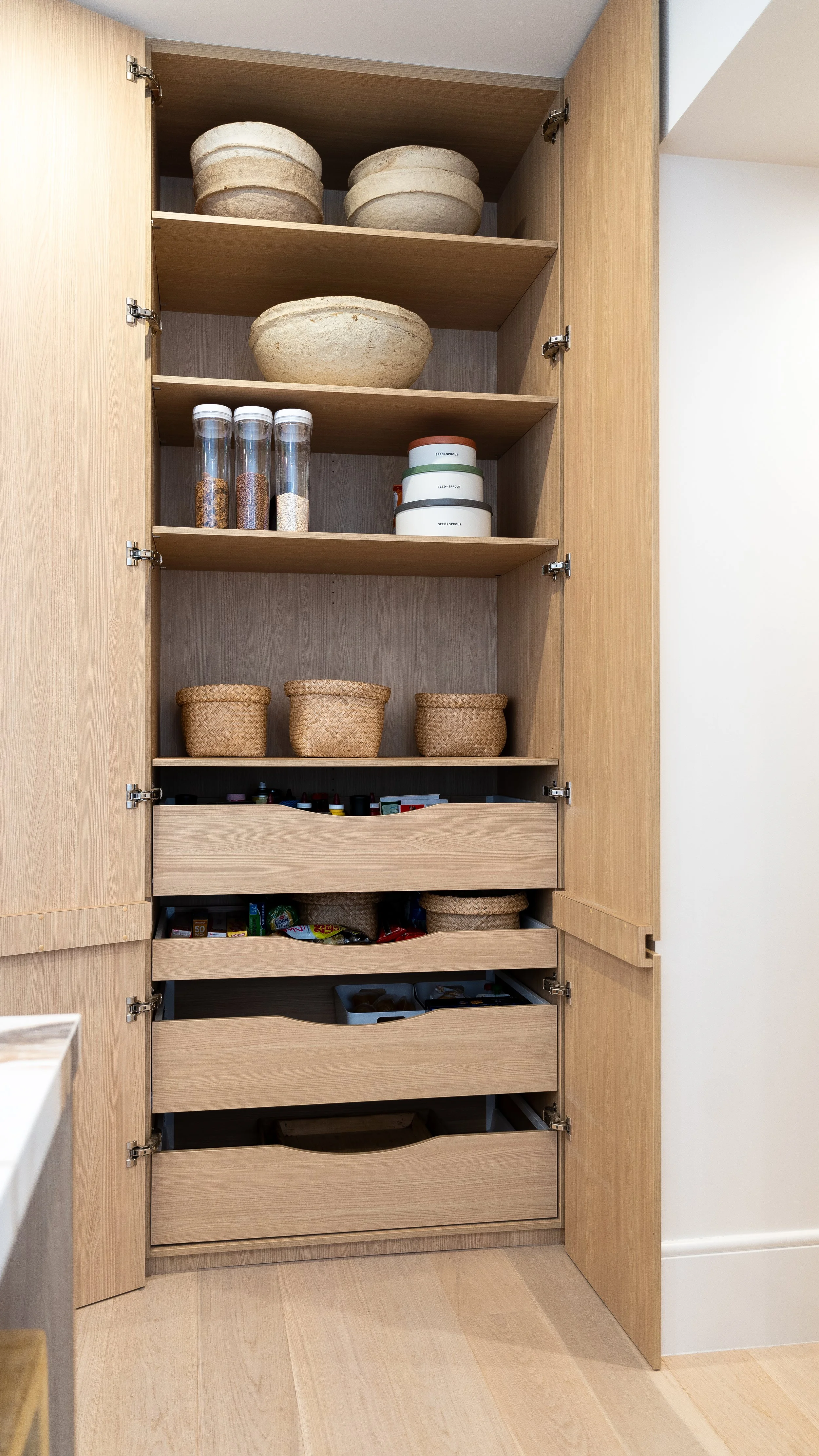 Open wooden pantry cabinet with shelves containing bowls, jars with grains or spices, and storage containers.