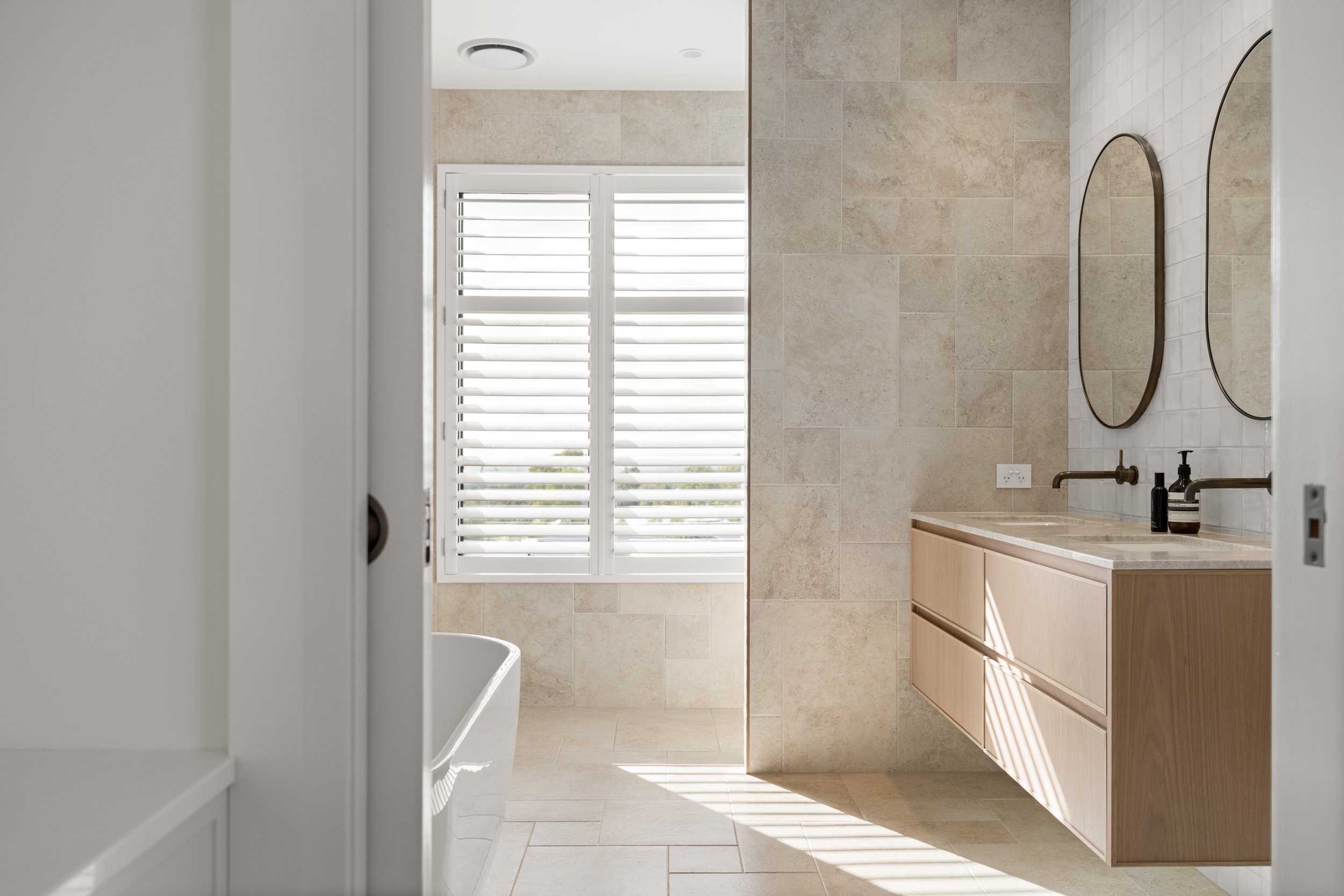 A modern bathroom with a large window with white plantation shutters, beige tiled walls, a wooden vanity with a marble countertop, a mirror, and sink accessories.
