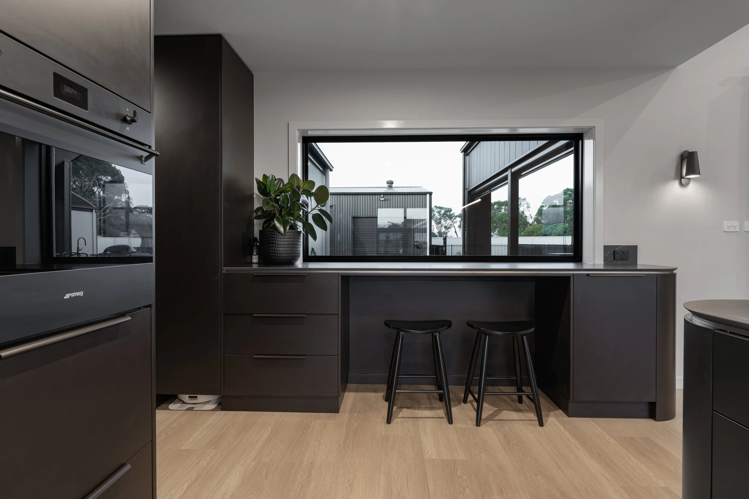 Modern kitchen with black cabinetry, large window, potted plant, and two black stools.