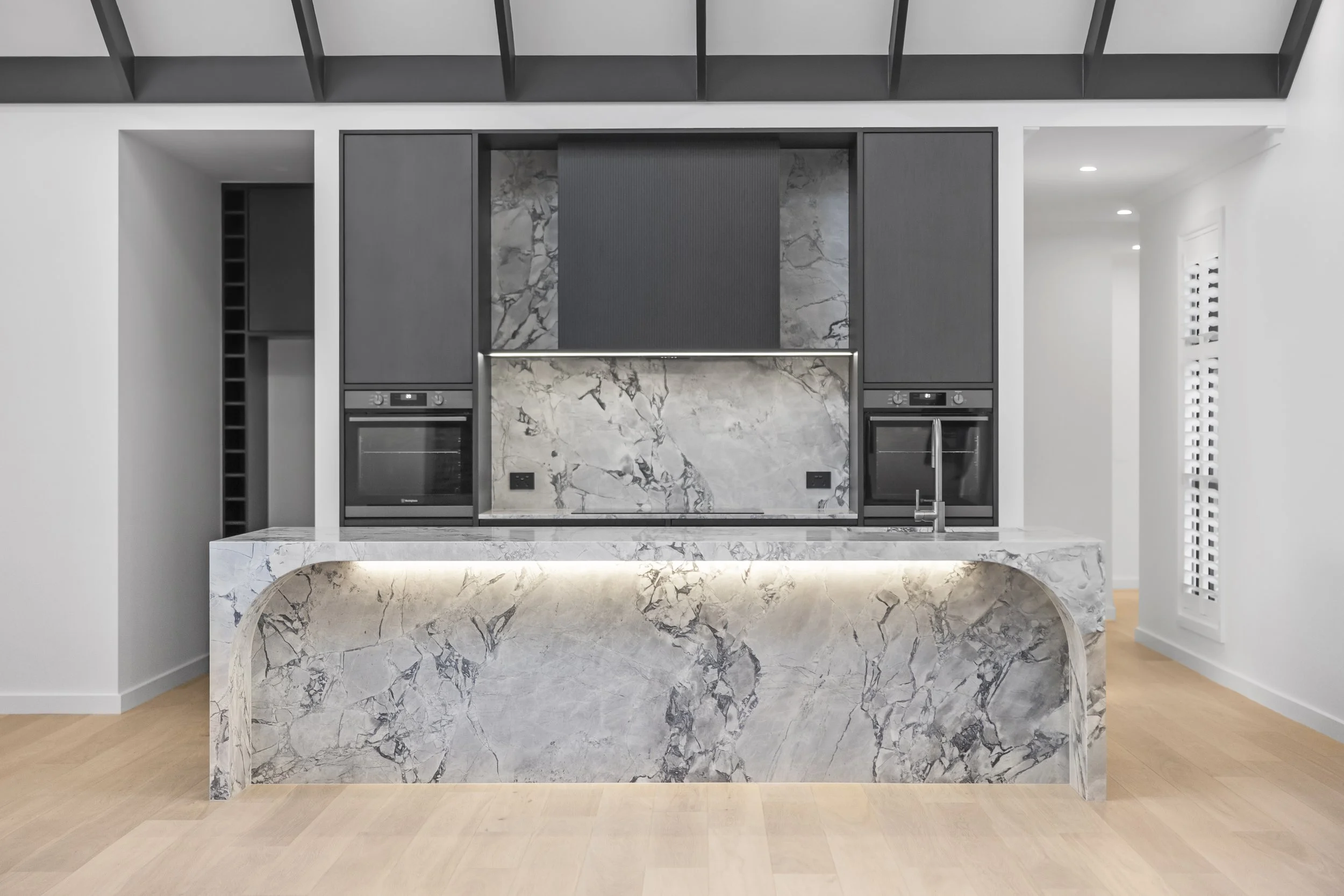 Modern kitchen with gray cabinetry, marble backsplash and island, built-in oven, and a faucet, with light wood flooring and white walls.