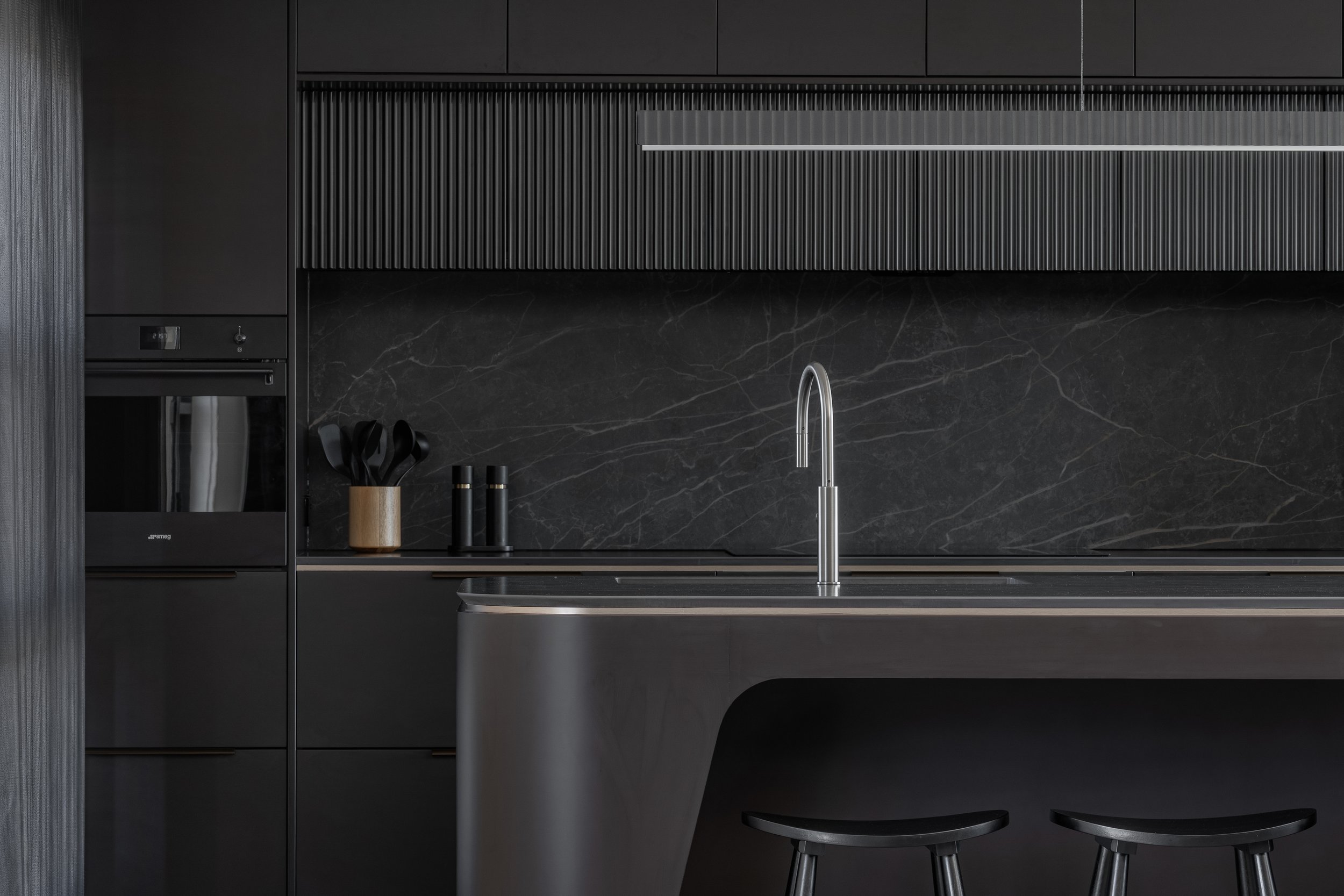 A modern kitchen with black cabinetry, a marble backsplash, a stainless steel faucet, a built-in oven, and black bar stools.