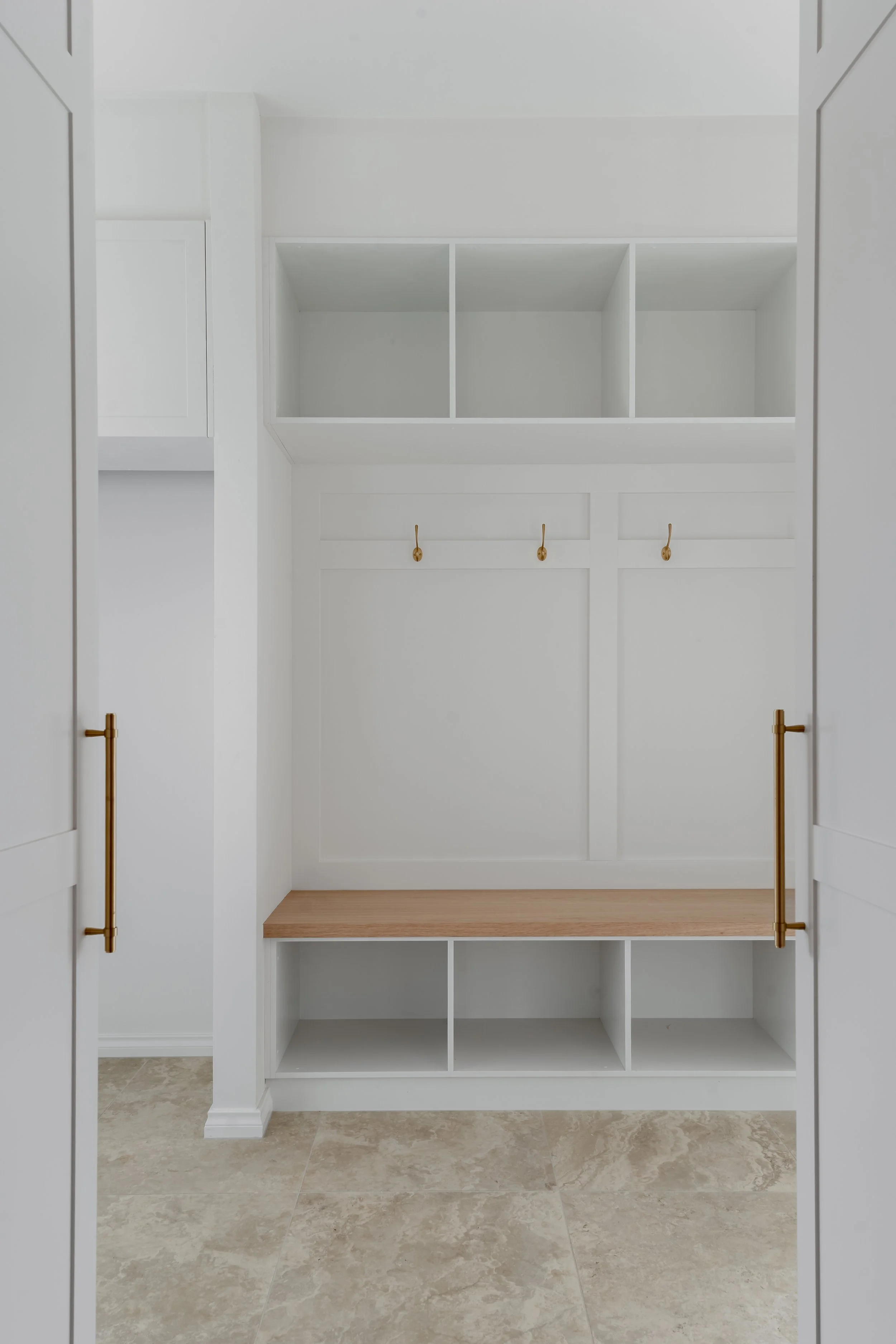 Empty white built-in closet with open doors, gold handles, and a wooden shelf, with a marble floor.
