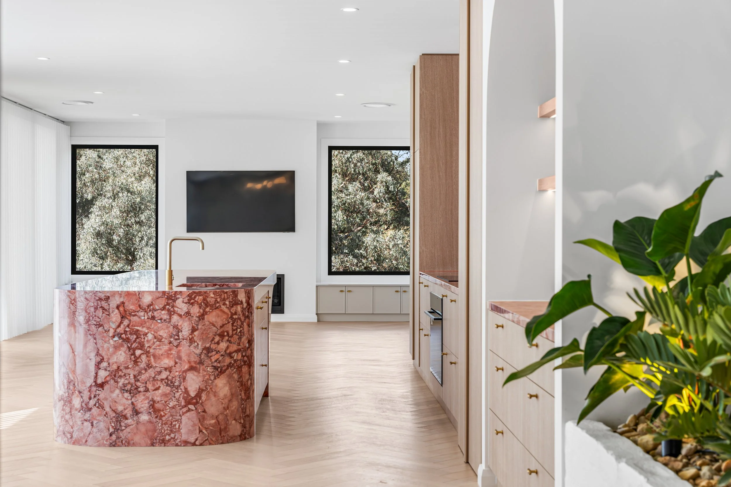 Modern kitchen with white walls, light wood floors, and a pink marble island with a gold faucet. Large windows show trees outside. Shelves and cabinets are visible, and a potted plant is on the right.