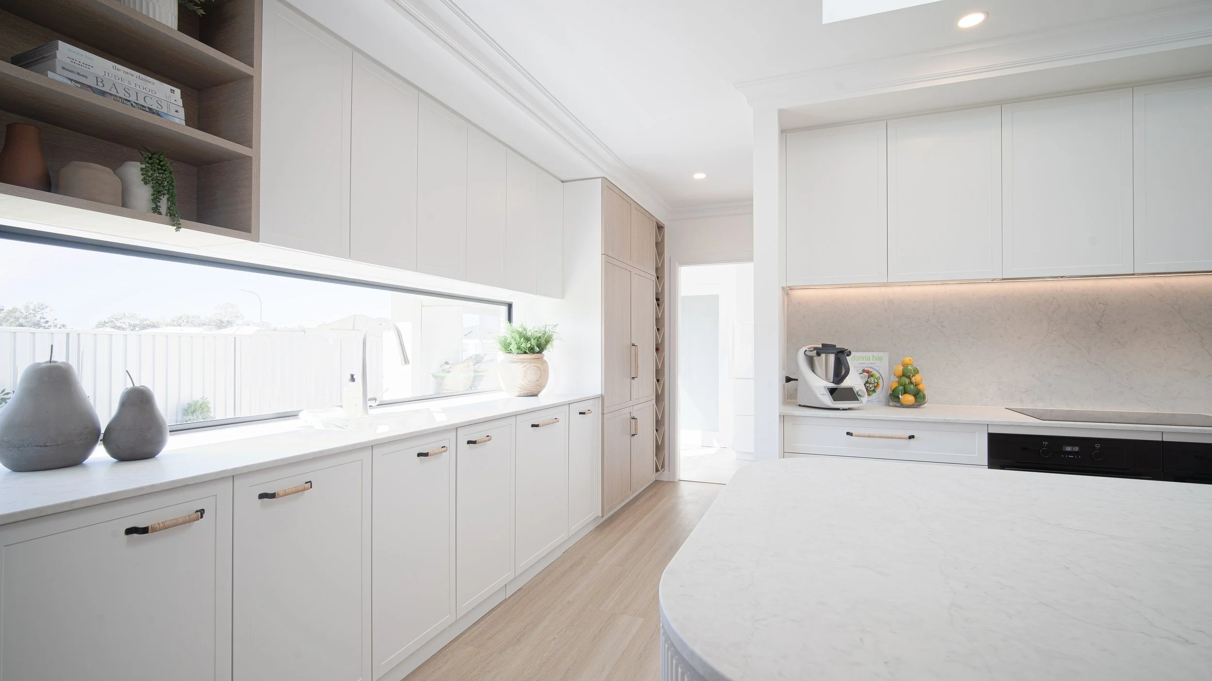 A bright modern kitchen with white cabinets, a marble countertop, and open shelving with decorative vases and books. A window above the sink lets in natural light, and there are appliances including a food processor and a bowl of lemons.