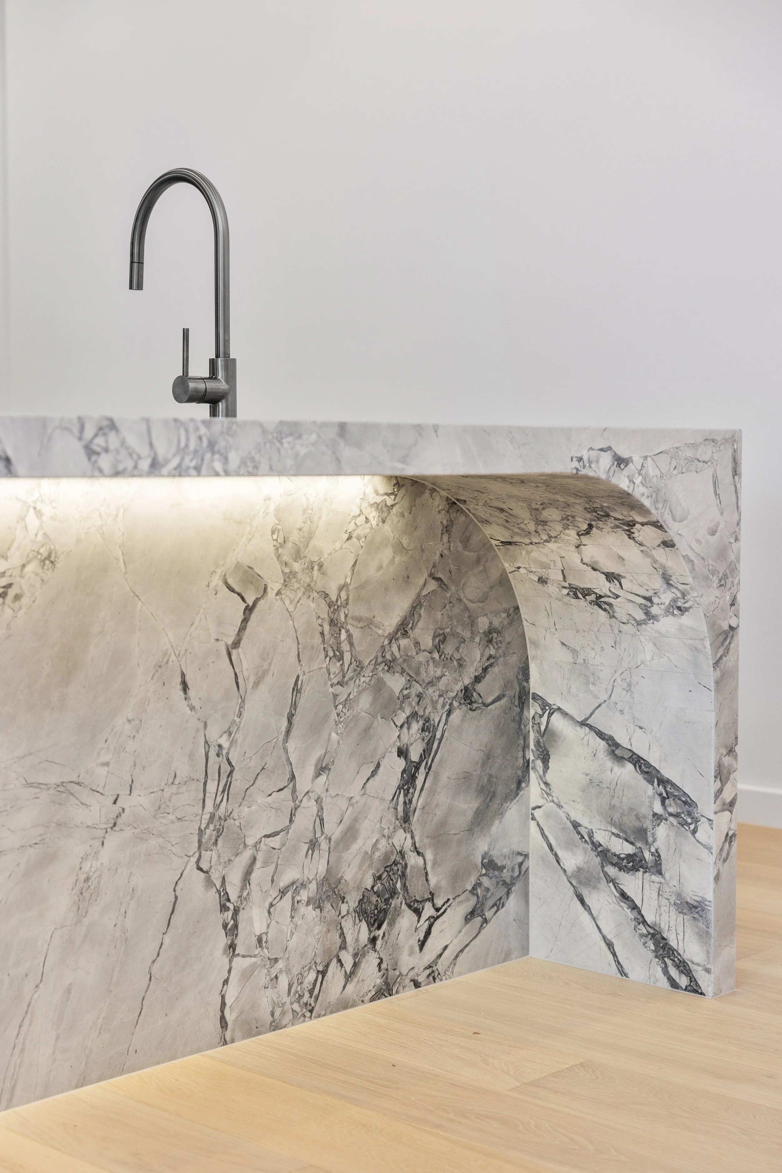 Close-up of a modern kitchen sink with a stainless steel faucet, set against a white marble countertop and splash guard, with a light wood floor in the background.