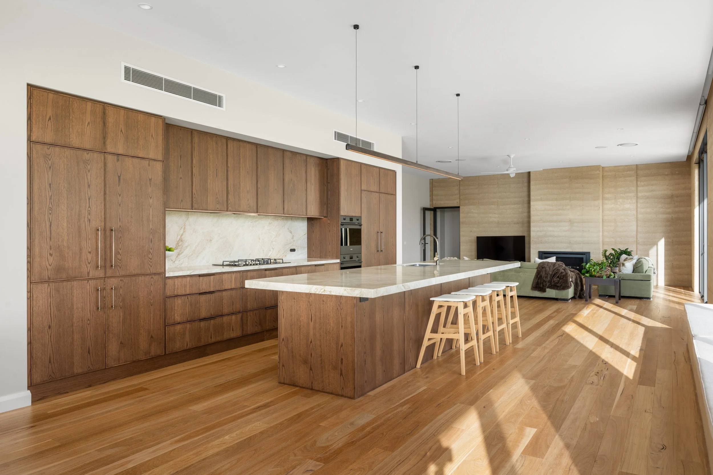 Open-concept kitchen and living area with wooden cabinetry, a large marble island, and a seating area with a sofa and plants, illuminated by natural light from large windows.